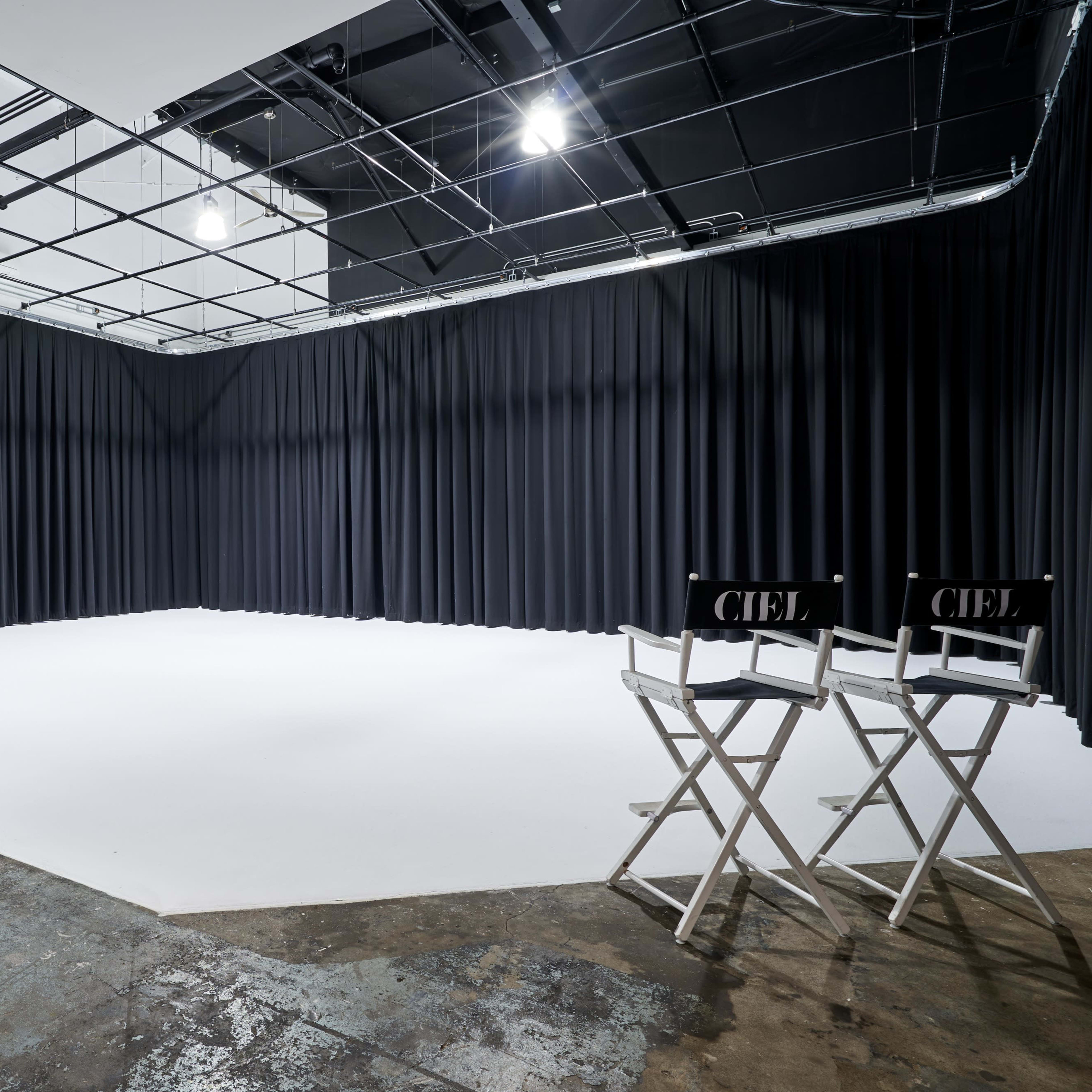 A spacious, empty studio with black curtains lining the walls and two director's chairs positioned on a white floor.