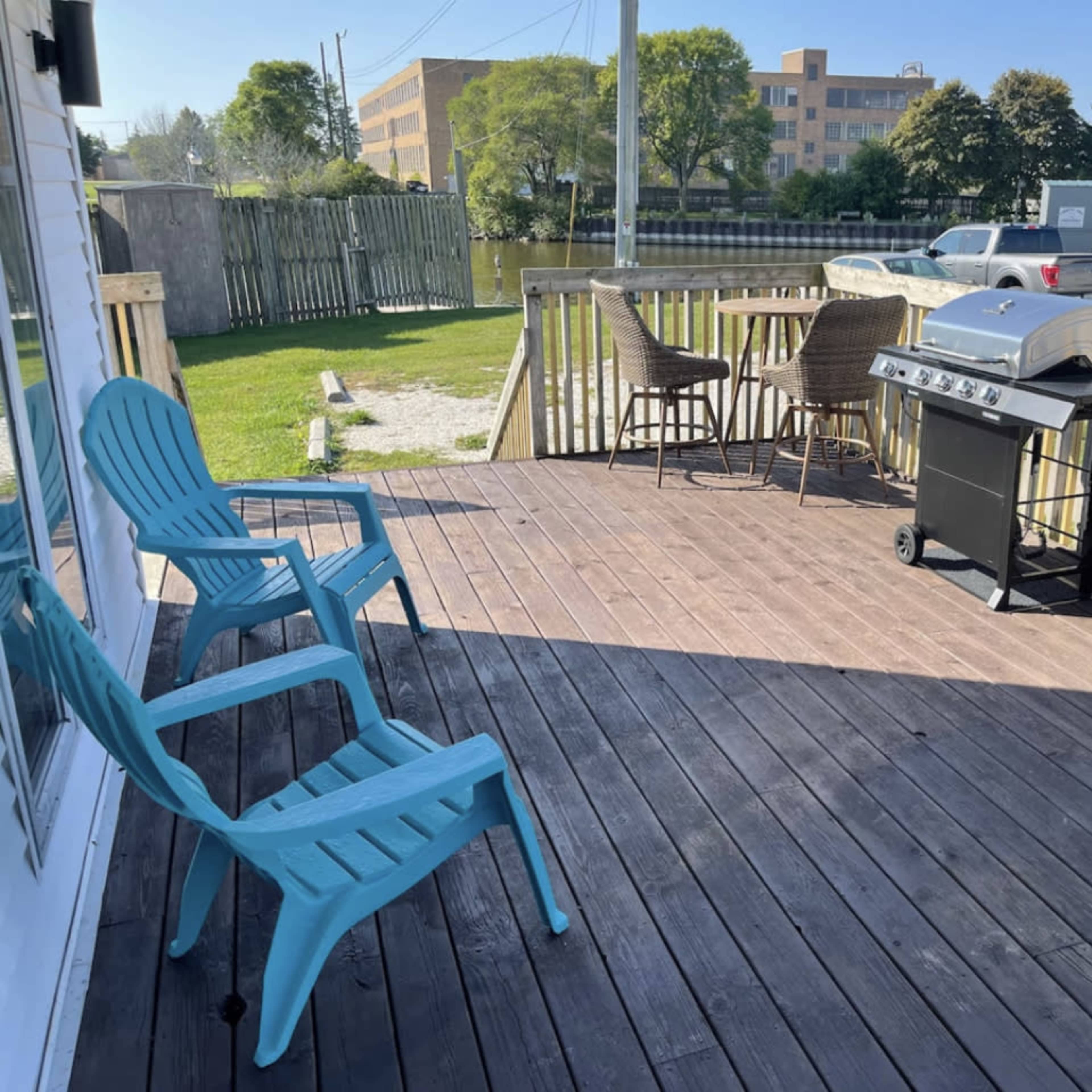 A wooden deck with blue chairs, a grill, and a view of a grassy area and buildings in the background.