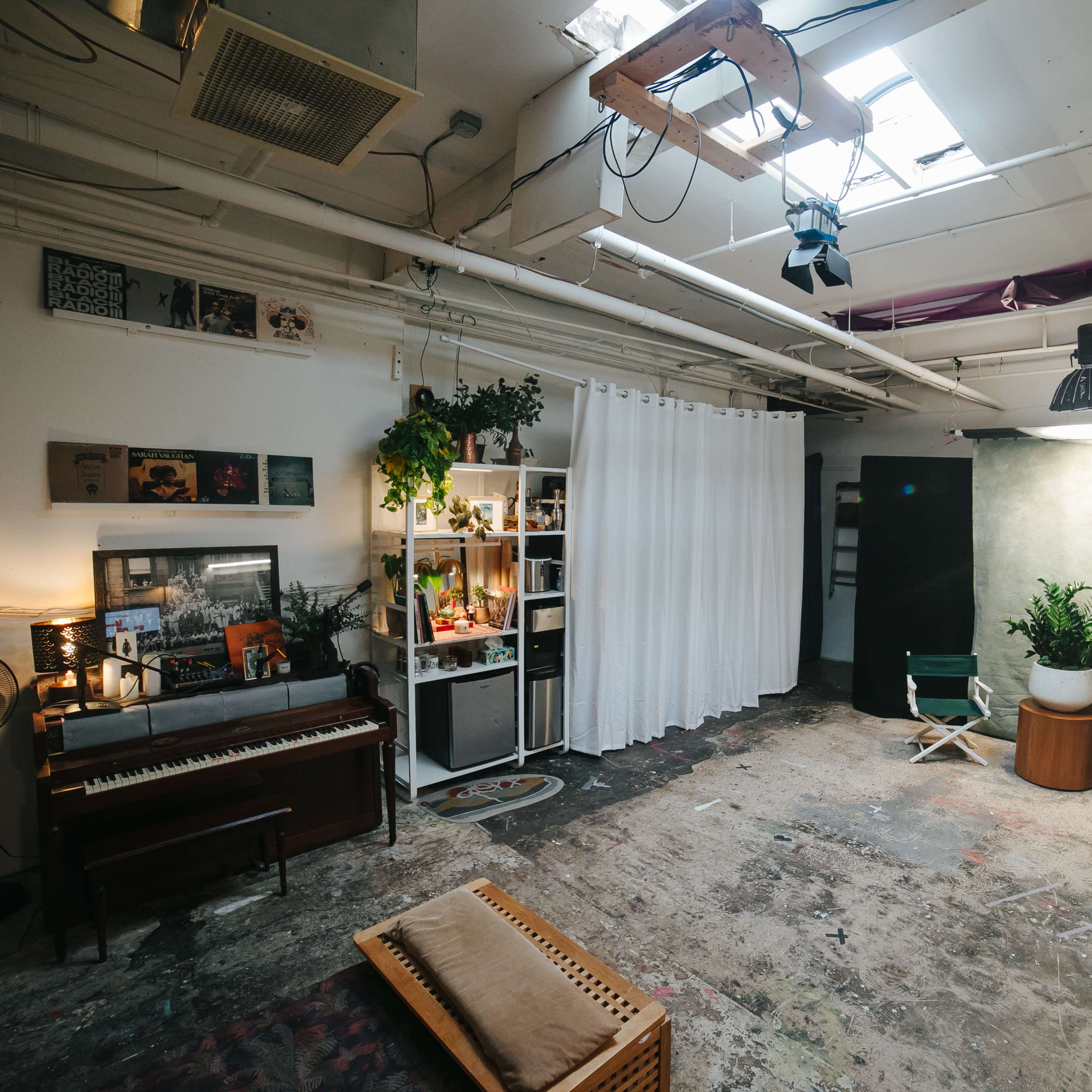 An industrial-style studio space featuring a piano, a curtain divider, plants, and various lighting fixtures.
