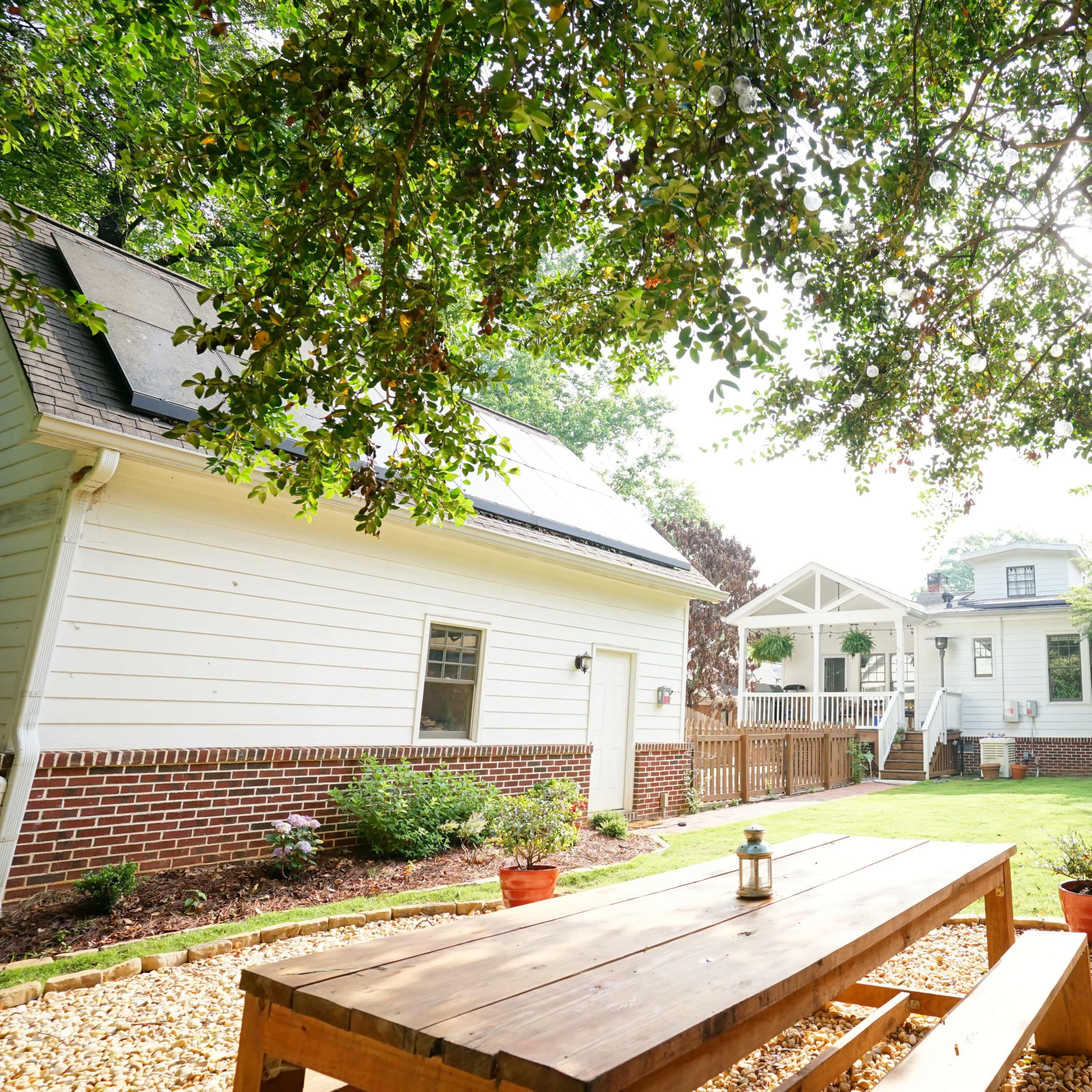 A wooden table sits in a landscaped backyard surrounded by trees and a wooden fence, with a house and deck in the background.