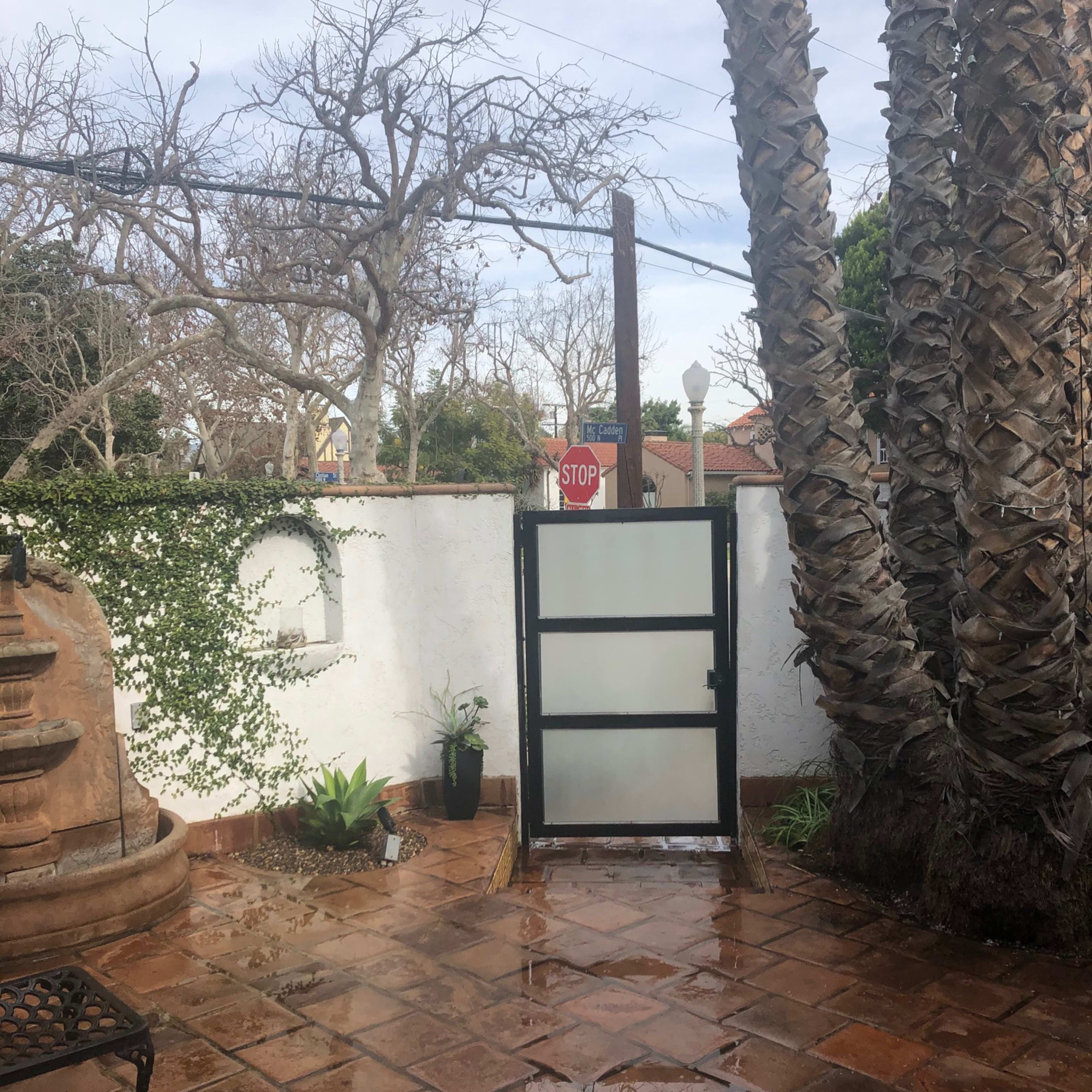 A courtyard with palm trees, a stop sign, and a partially open glass door, surrounded by a white wall and tiled floor.