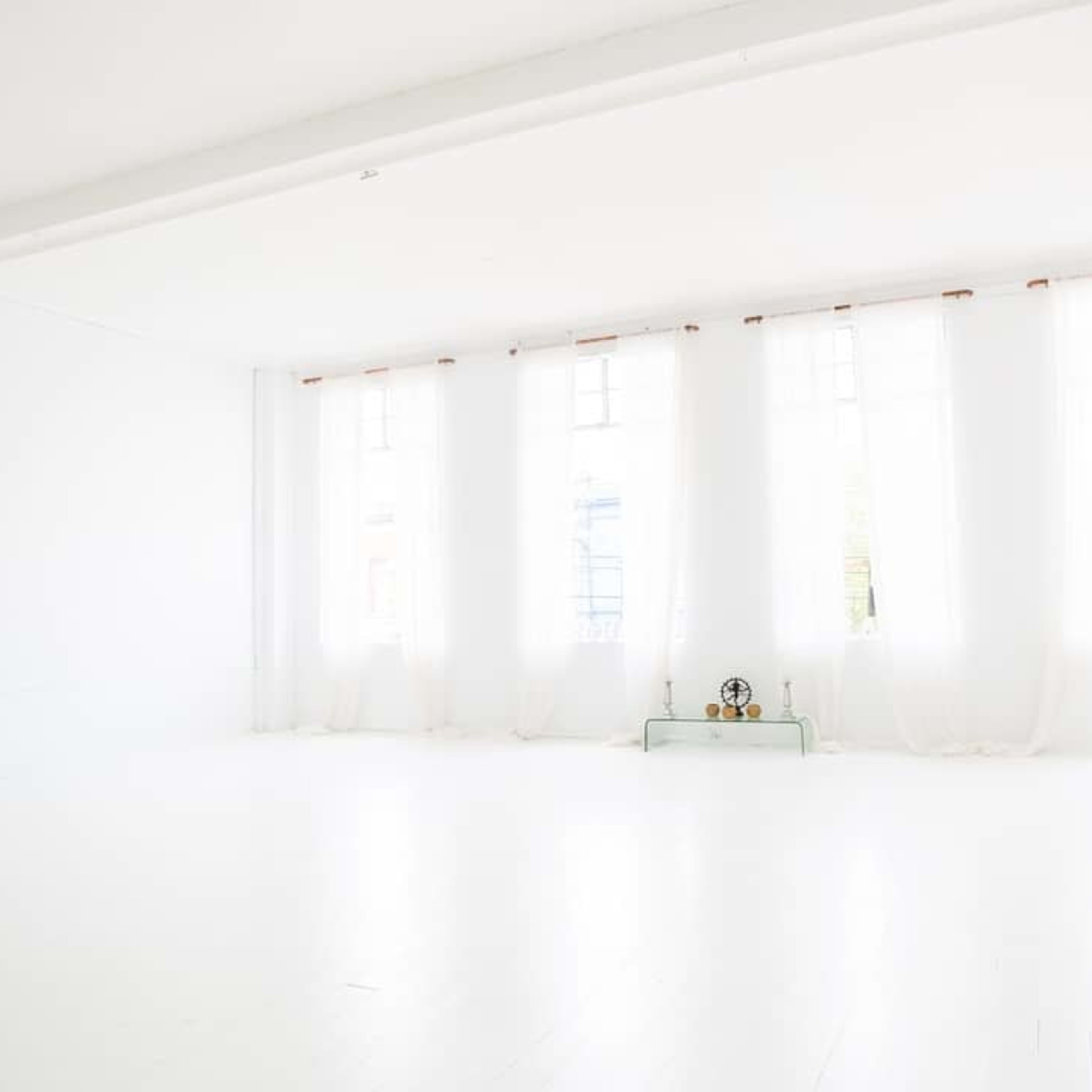 A spacious, brightly lit room features large windows with sheer curtains and a minimalist glass shelf displaying a few decorative items.