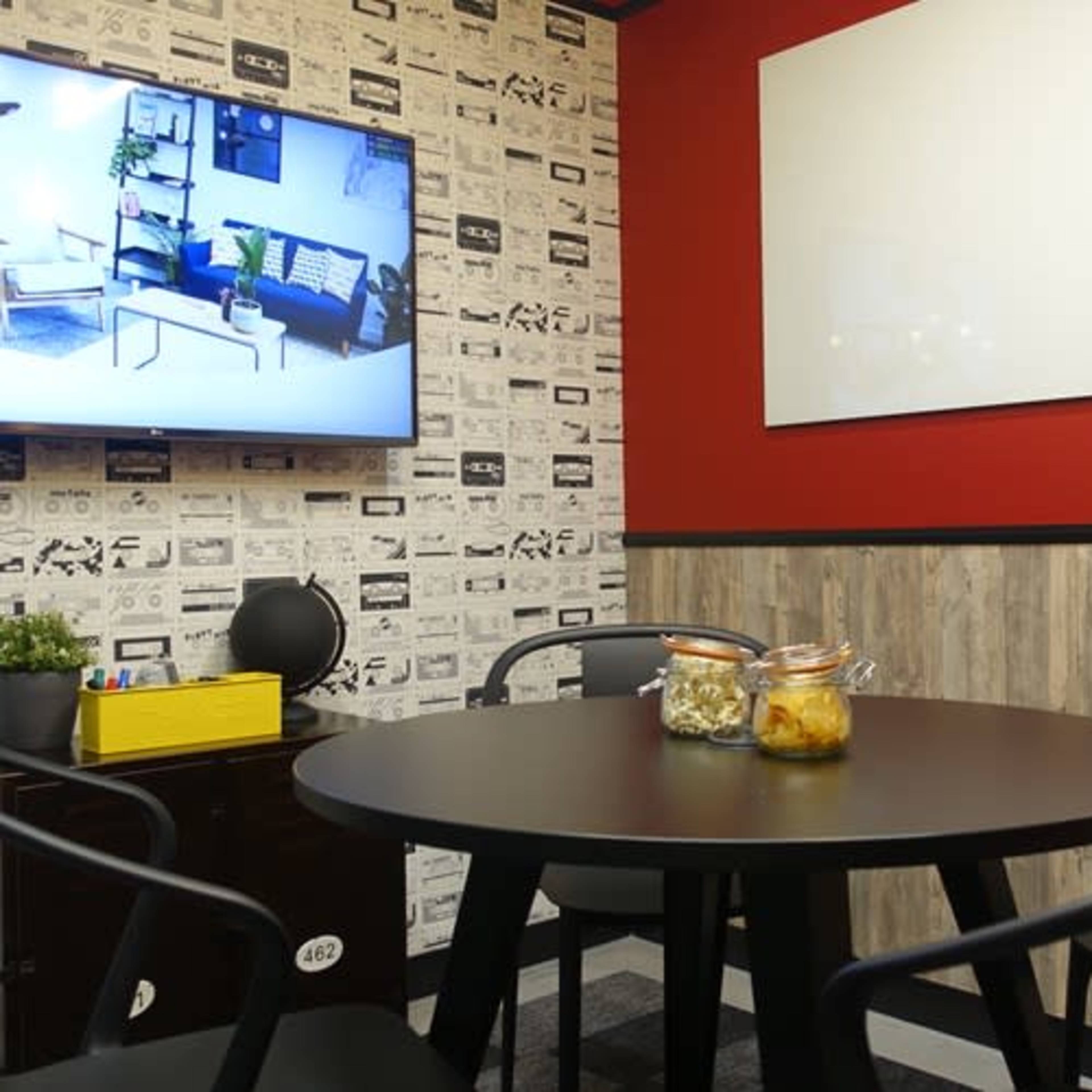 A small meeting room featuring a round black table with jars of snacks, a TV on the wall displaying a living room, and a whiteboard in a brightly colored space with patterned wallpaper.