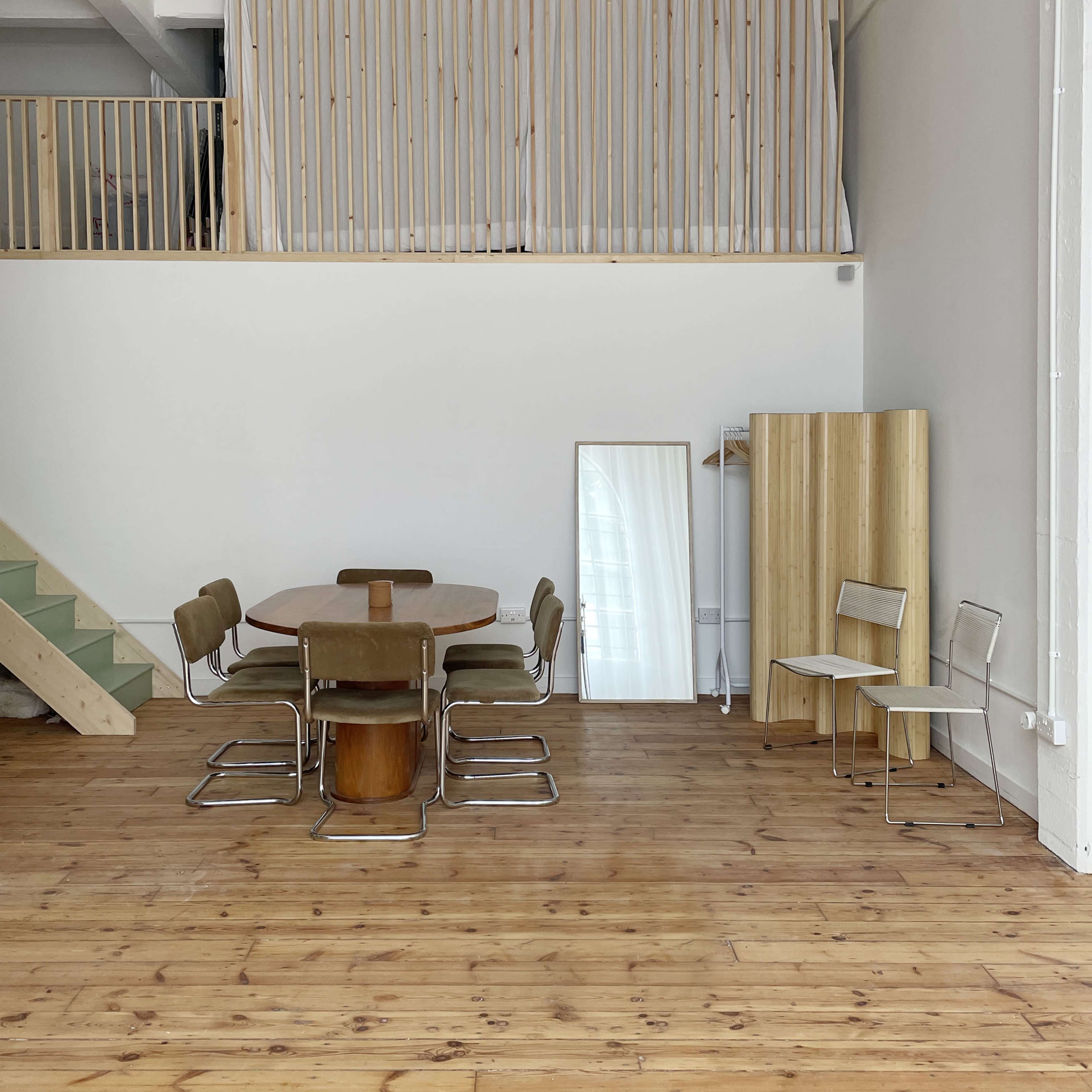 A minimalist interior space features a wooden dining table surrounded by metal chairs, with a staircase and wooden barriers in the background.