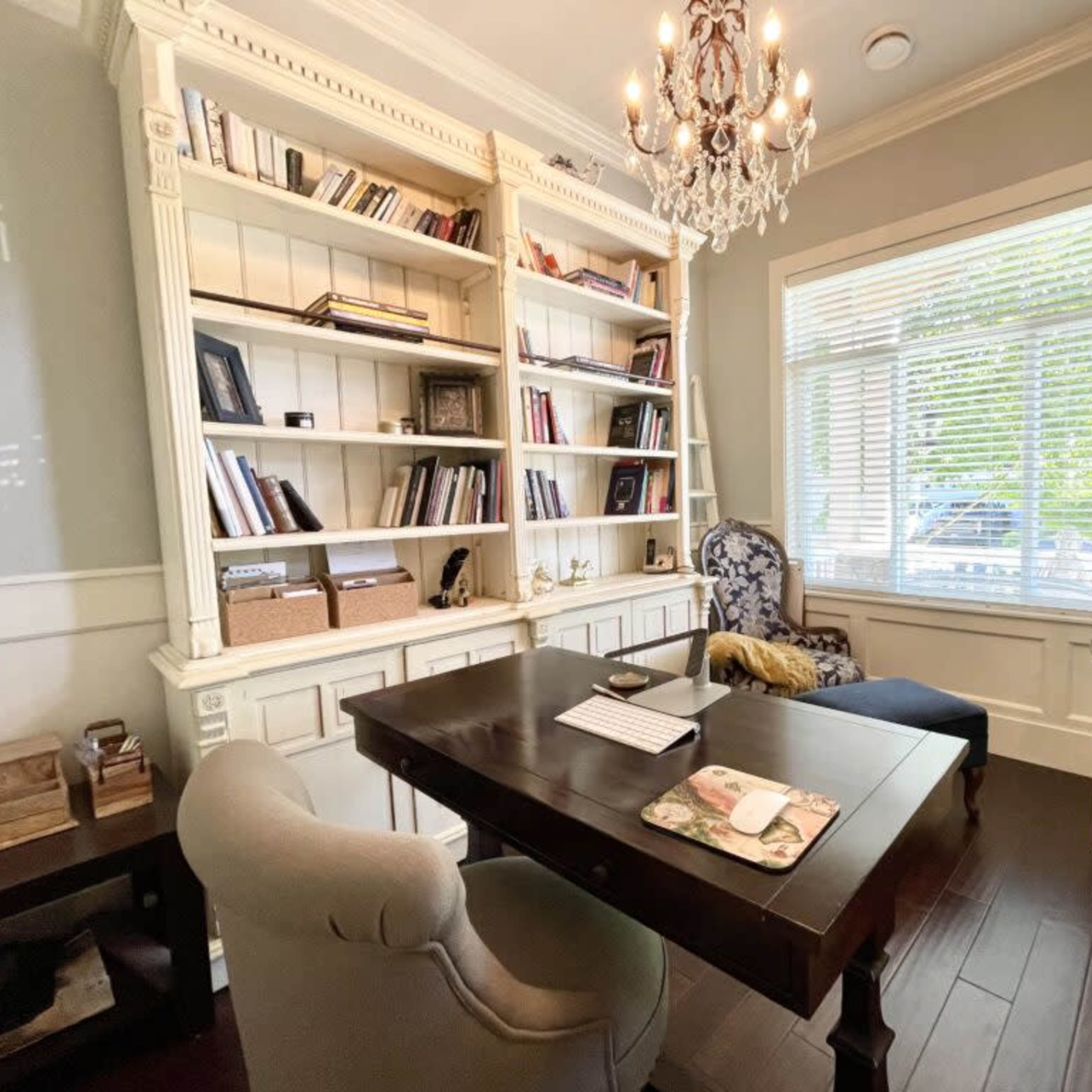 A well-organized home office featuring a dark wooden desk, an upholstered chair, a bookshelf filled with books, and a decorative chandelier.