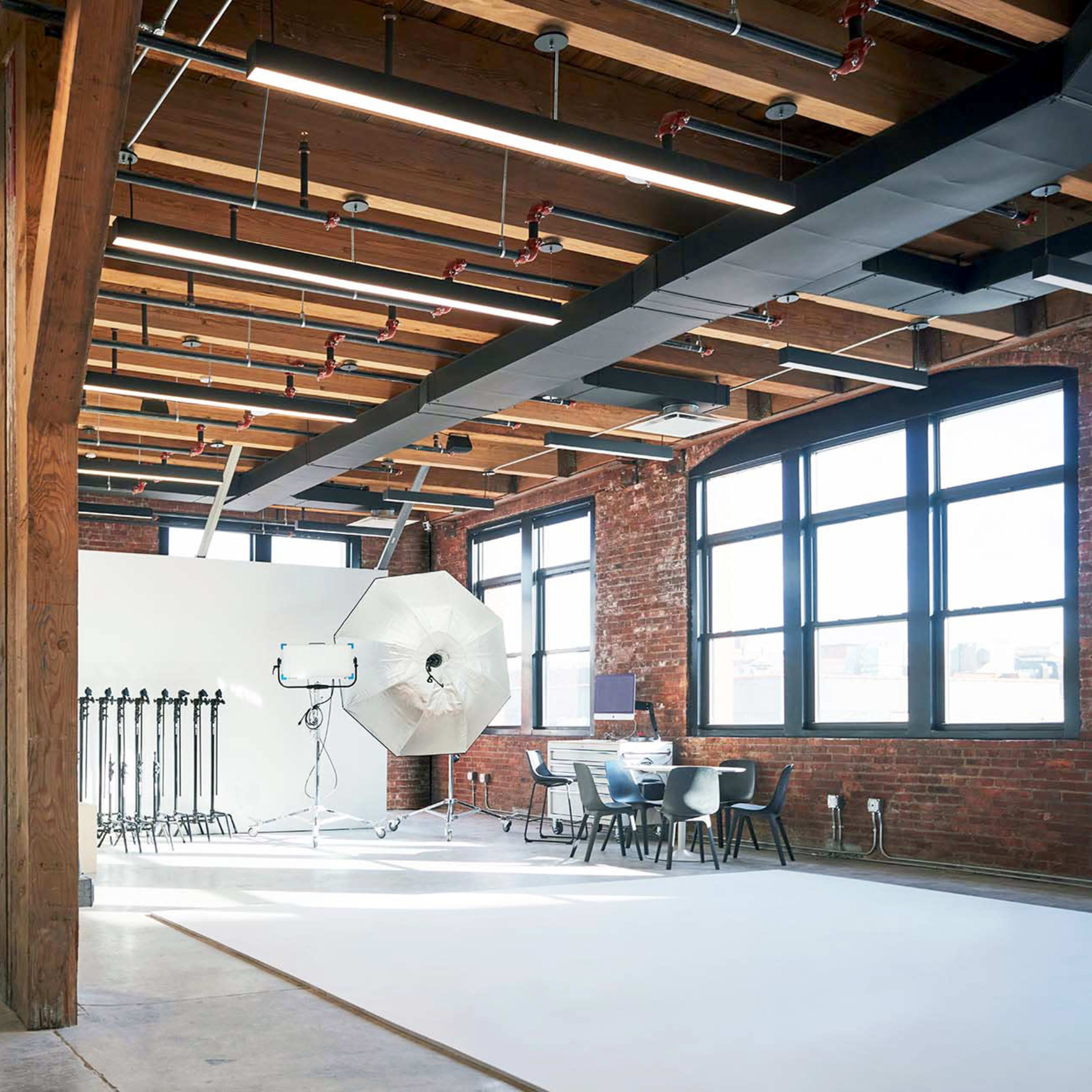 A spacious studio features exposed wooden beams, large windows, and photography equipment set up against a white wall.