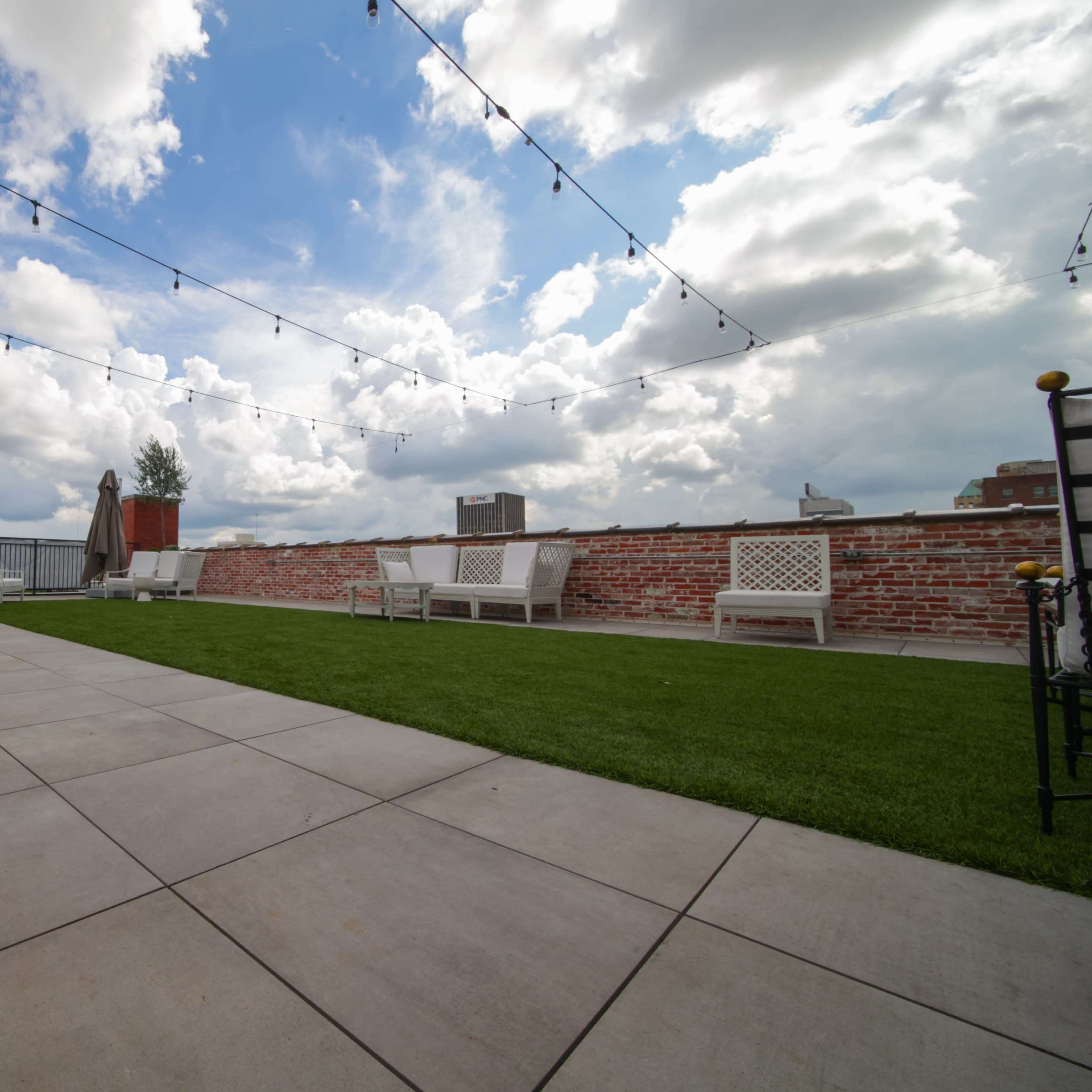 A rooftop terrace featuring lounge seating and string lights overhead, with a grassy area and a backdrop of cloudy skies.