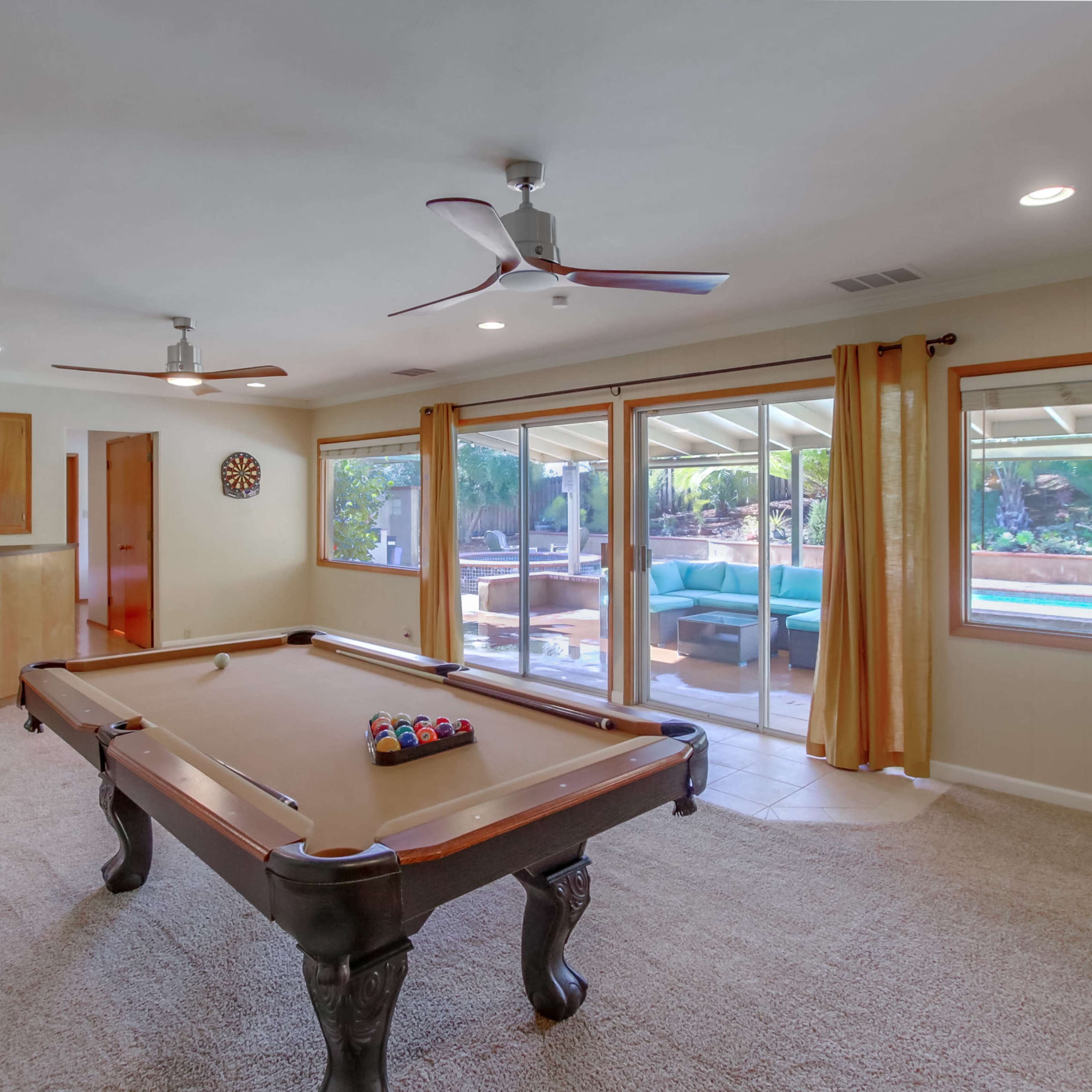 A billiards table is positioned in a well-lit room that features large windows overlooking a pool area.