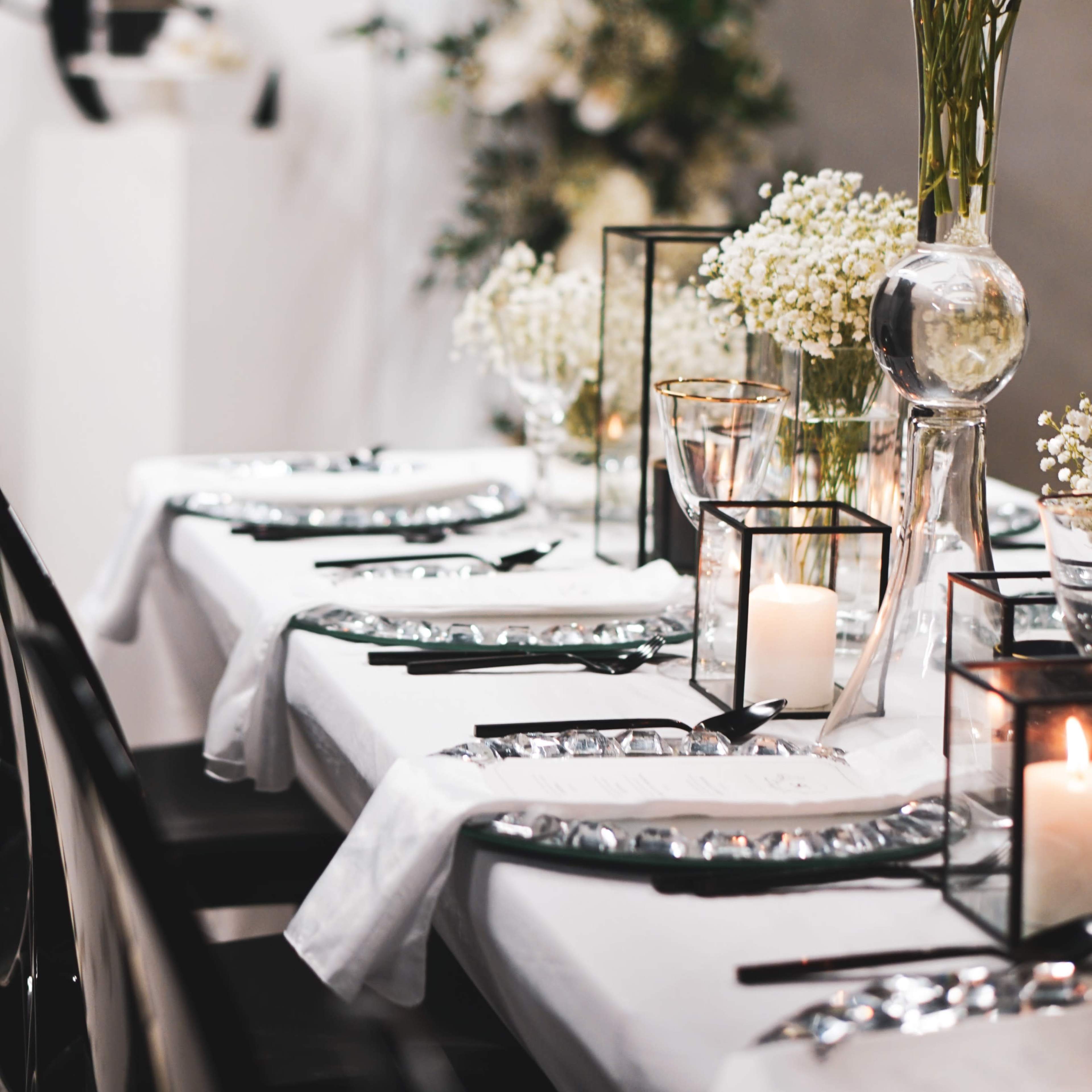 A elegantly set dining table features white tablecloths, glass vases with flowers, candles, and decorative accents in a styled venue.