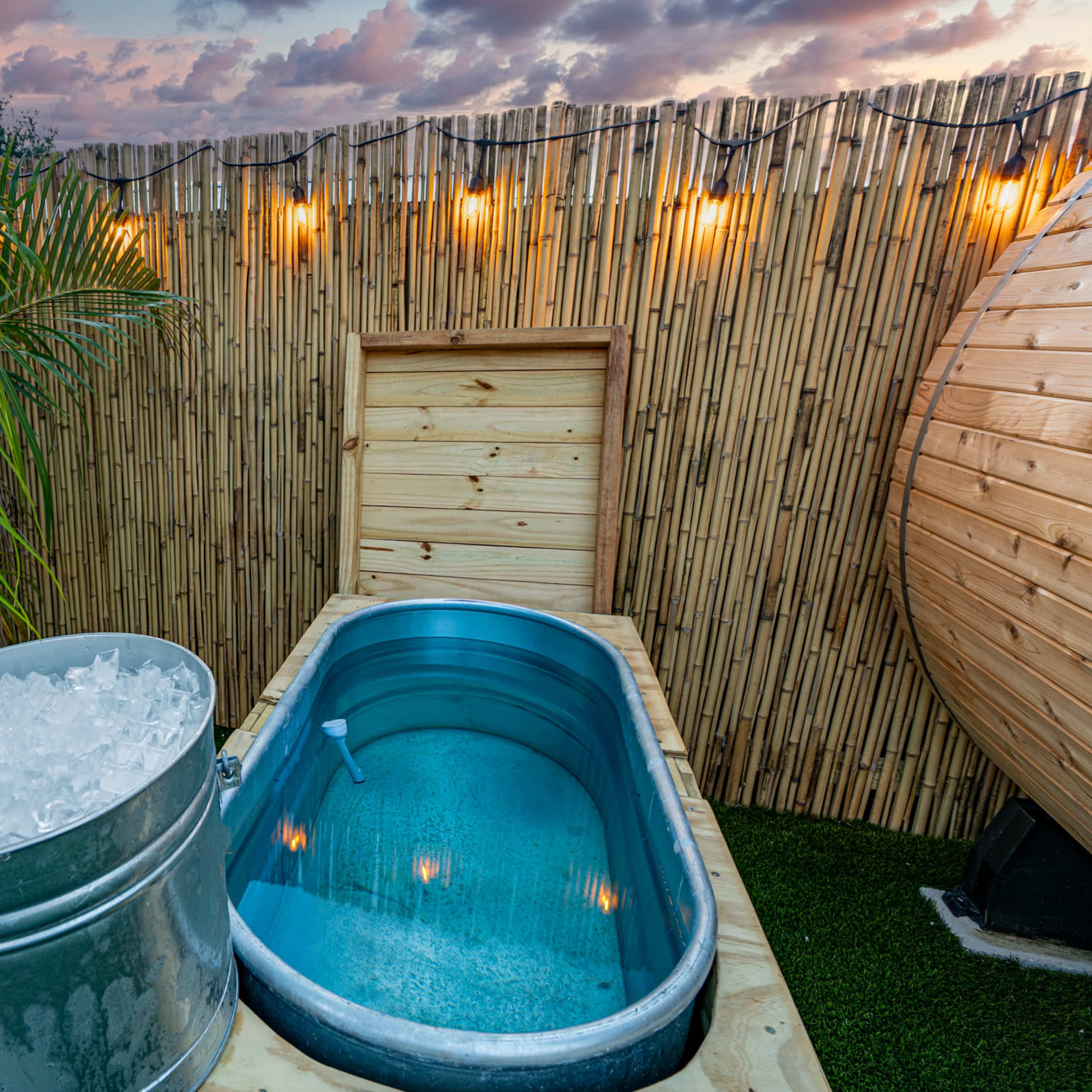 A wooden hot tub surrounded by a bamboo fence, with a bucket of ice beside it and a pink flamingo decoration in the corner.