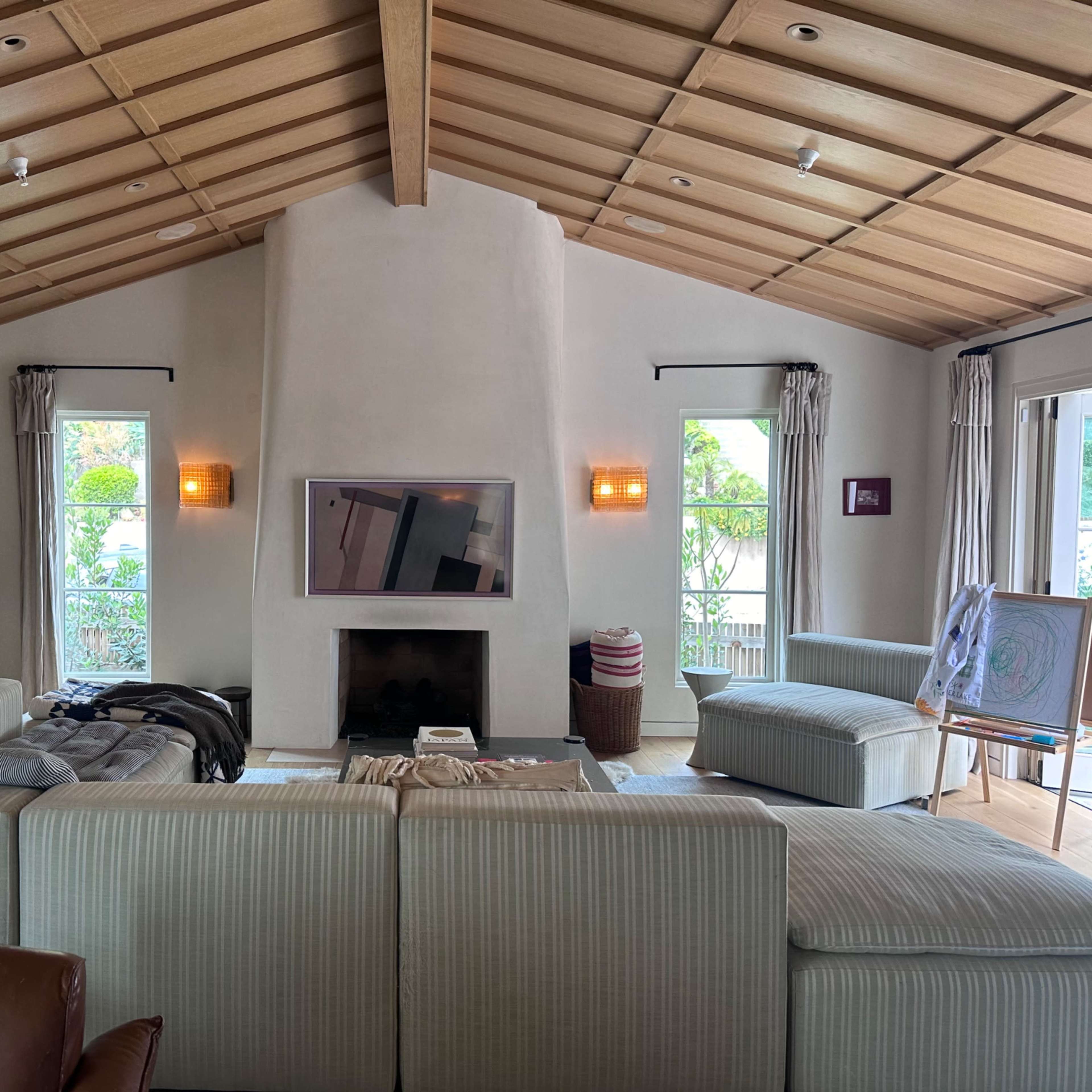 A spacious living room featuring a large sectional couch, a flat-screen TV mounted above a fireplace, and large windows providing views of greenery outside.