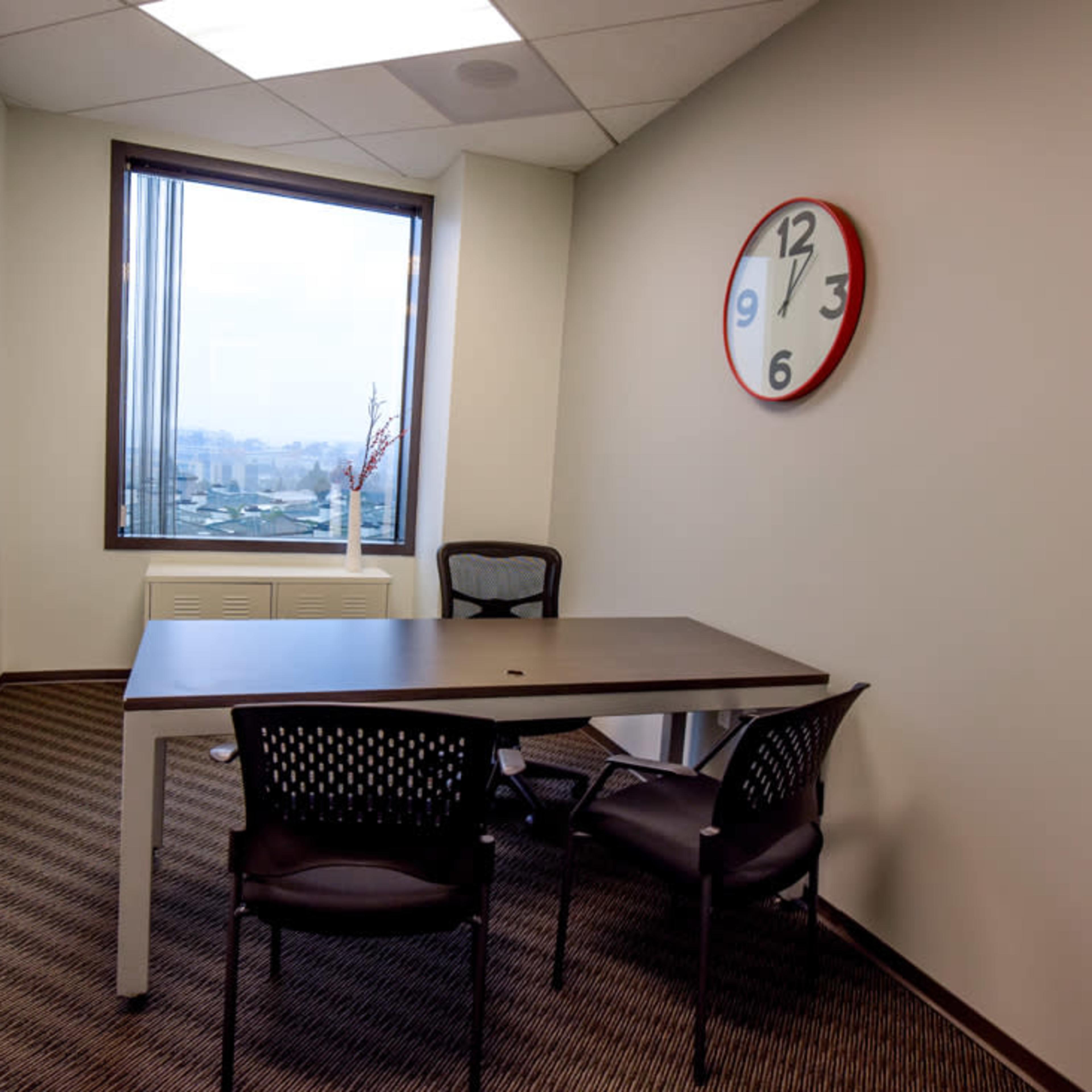 A small office meeting room featuring a table, two chairs, a wall clock, and a window with a view.