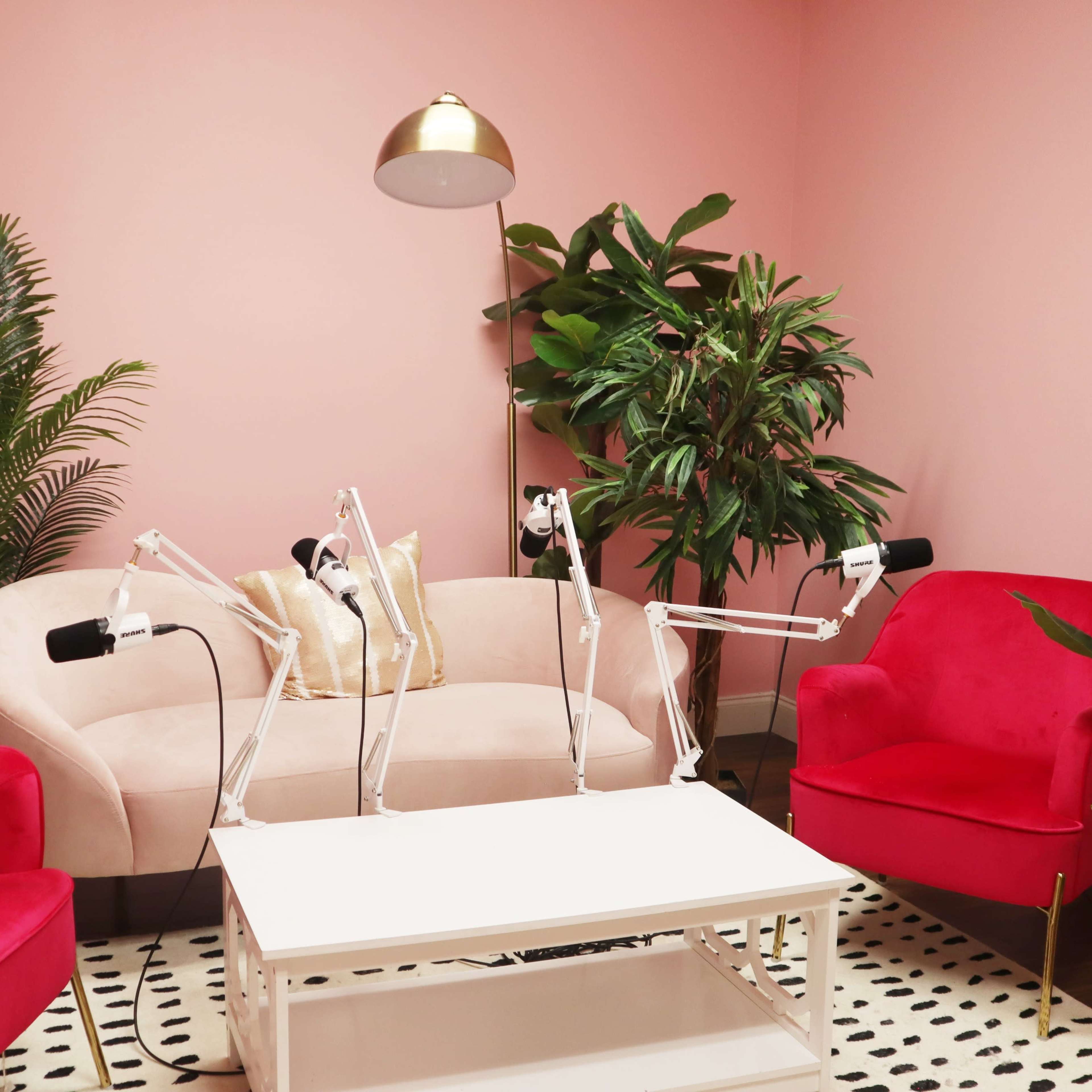 A modern seating area features a light pink sofa and two bright red armchairs, surrounded by greenery and a white coffee table with microphones for recording.