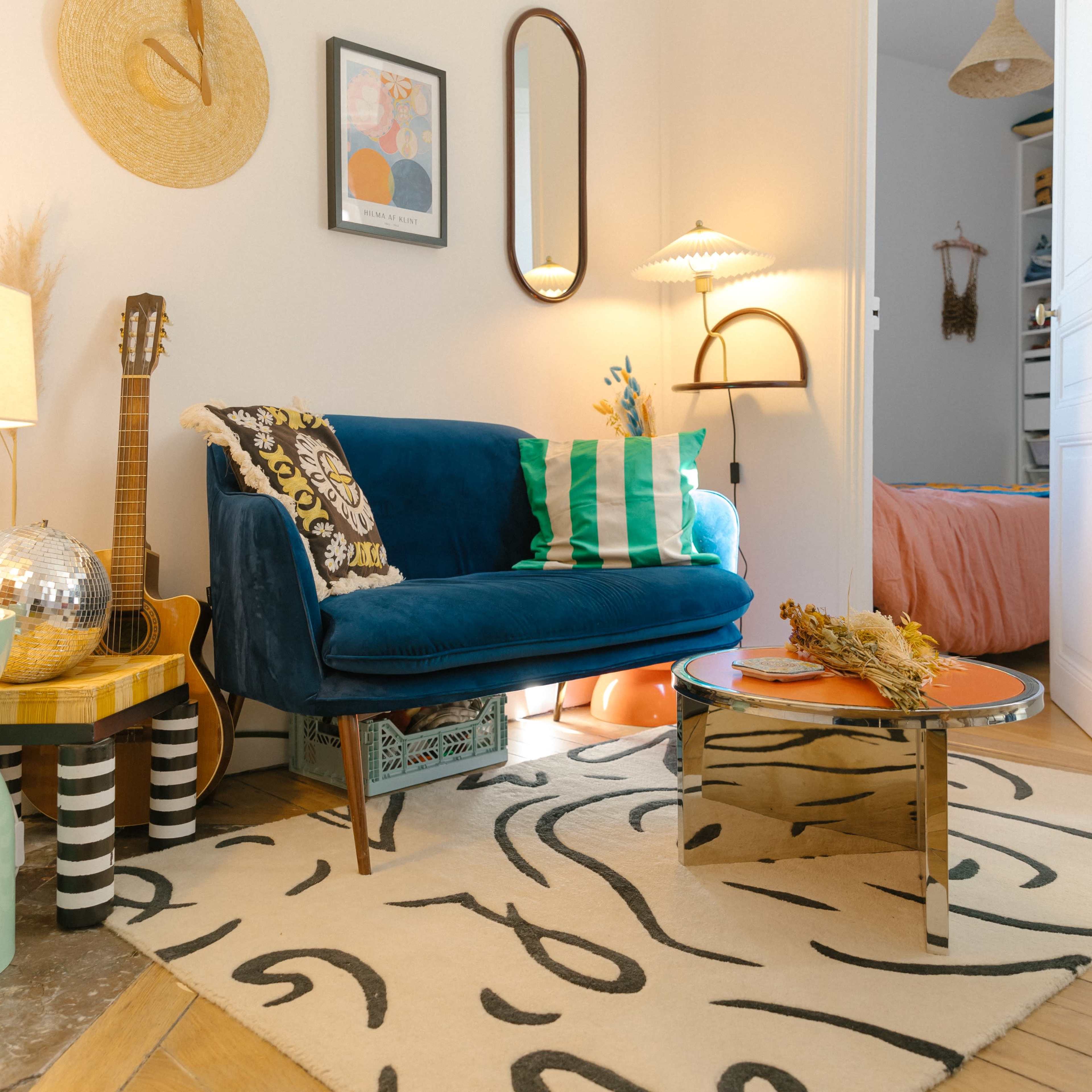 A cozy living room with a blue velvet sofa, a decorative rug, and various accessories, including a guitar, a lamp, and a round table.