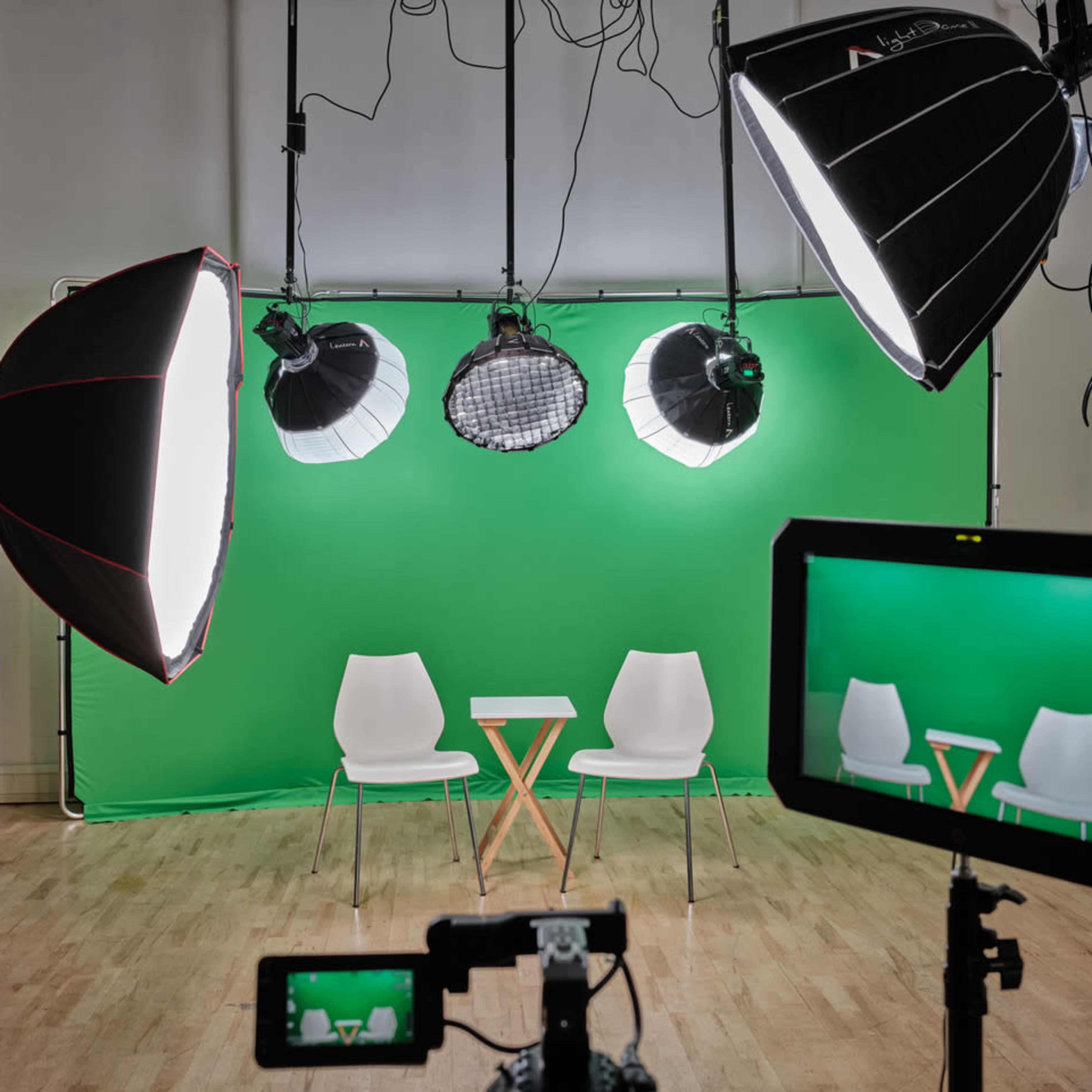 A studio setup features two white chairs facing a small table in front of a green backdrop, illuminated by five overhead lights and monitored by a camera.