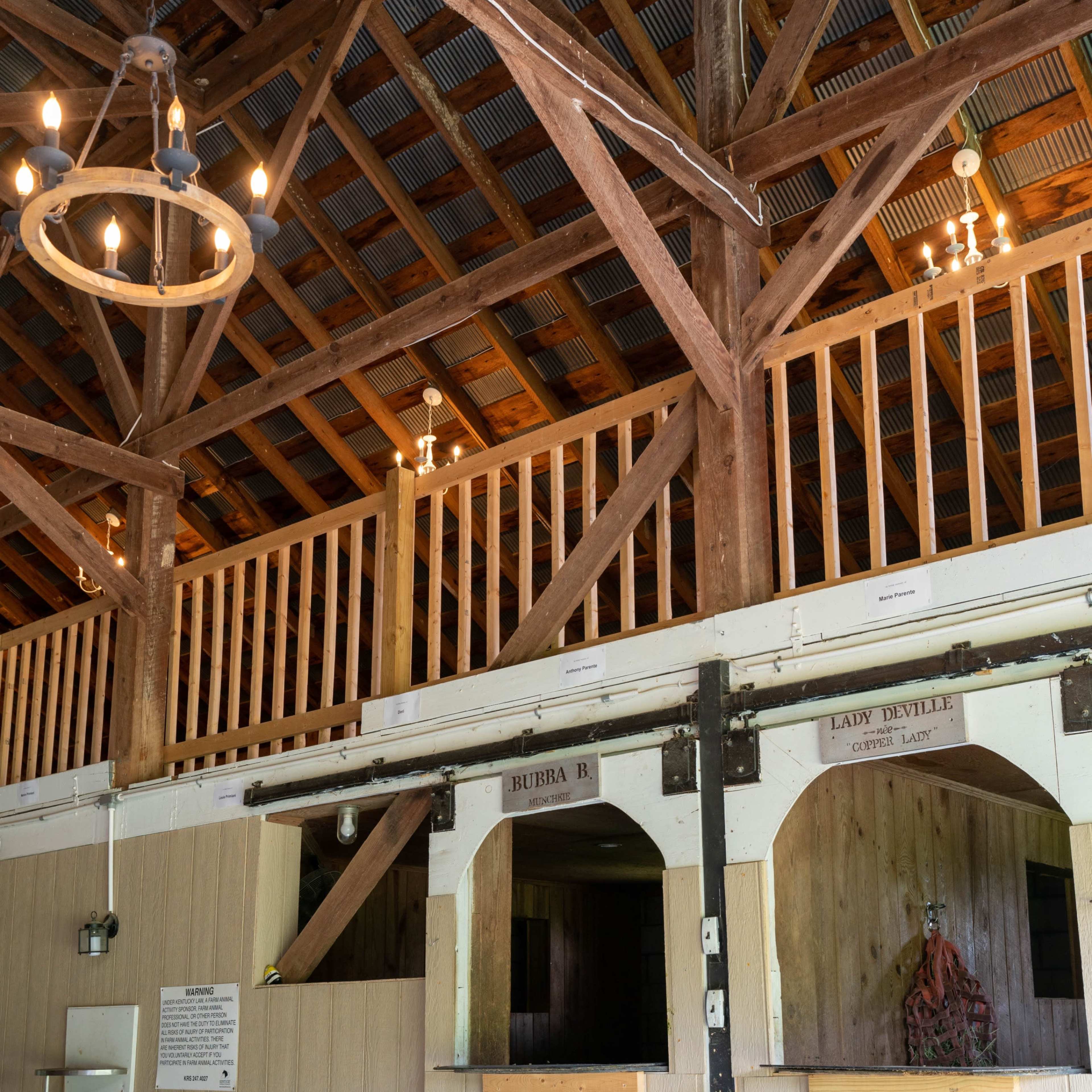 The interior of a barn featuring wooden beams, a second-story balcony, and a pair of horse stalls with names labeled above them.