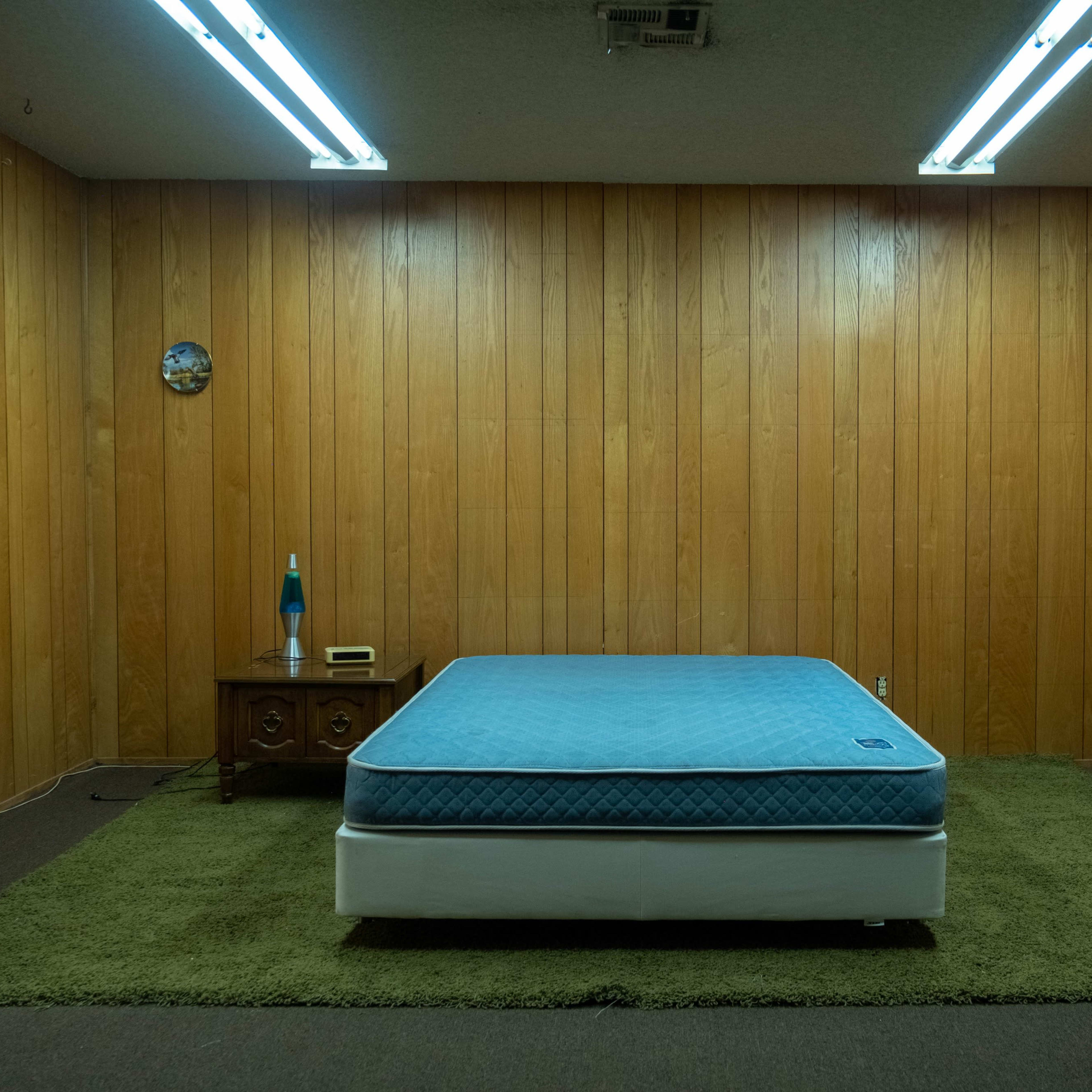 A sparsely furnished room with wood paneled walls, a blue mattress resting on a white base, a green carpet, a fan, and a dresser.