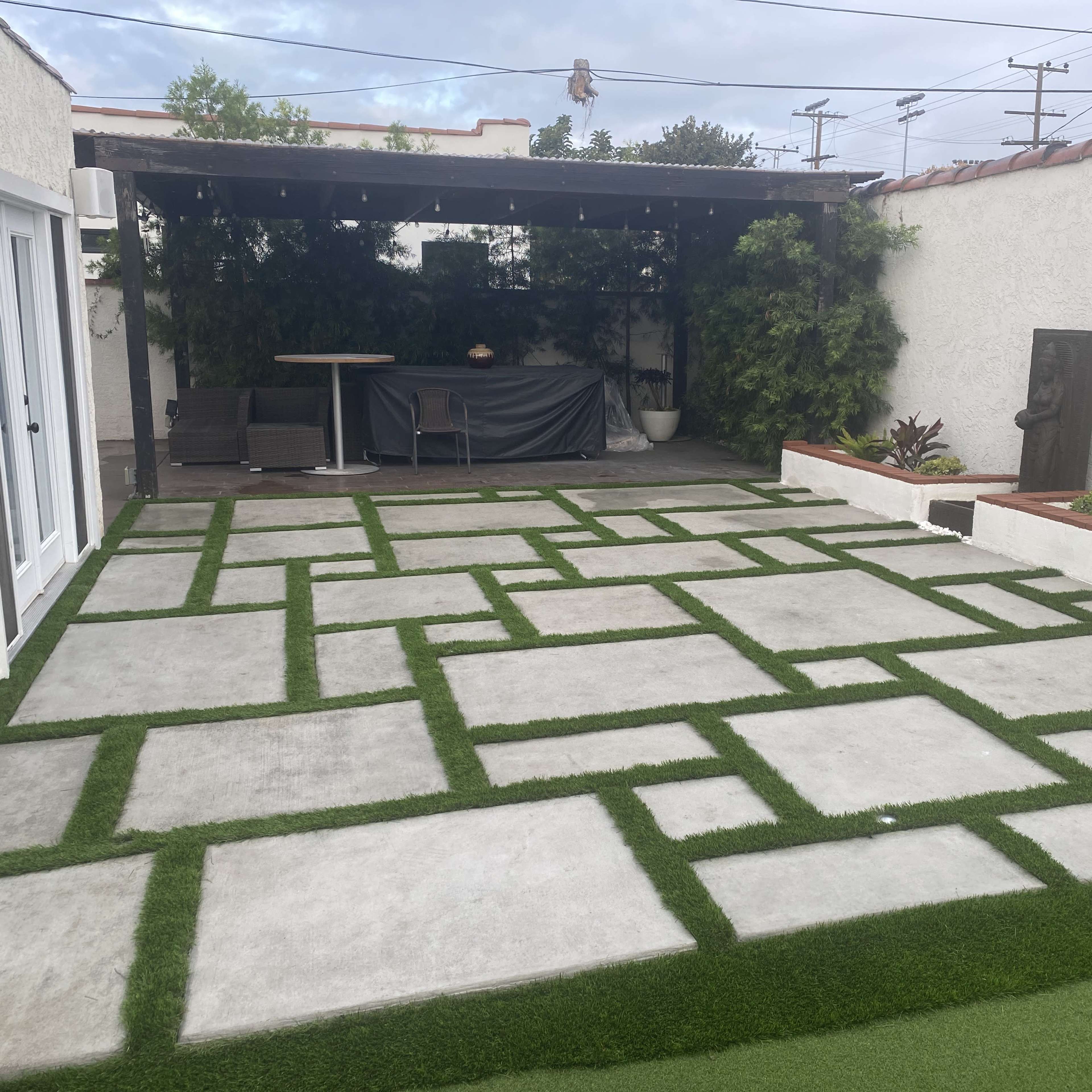 A rectangular patio with a geometric pattern of concrete slabs surrounded by artificial grass and a covered seating area in the background.