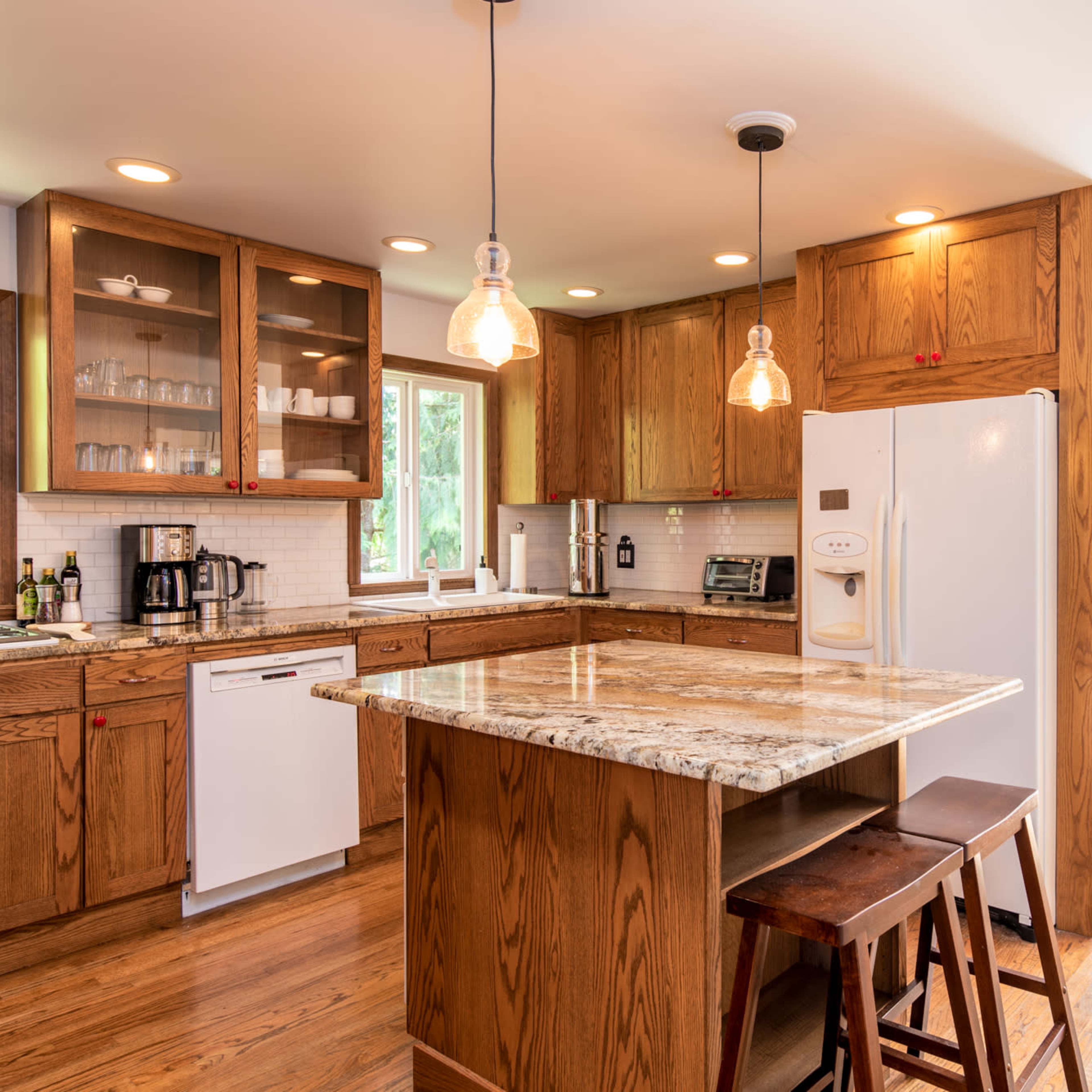 A modern kitchen features wooden cabinets, a central island with a granite countertop, and a white refrigerator, illuminated by pendant lights.