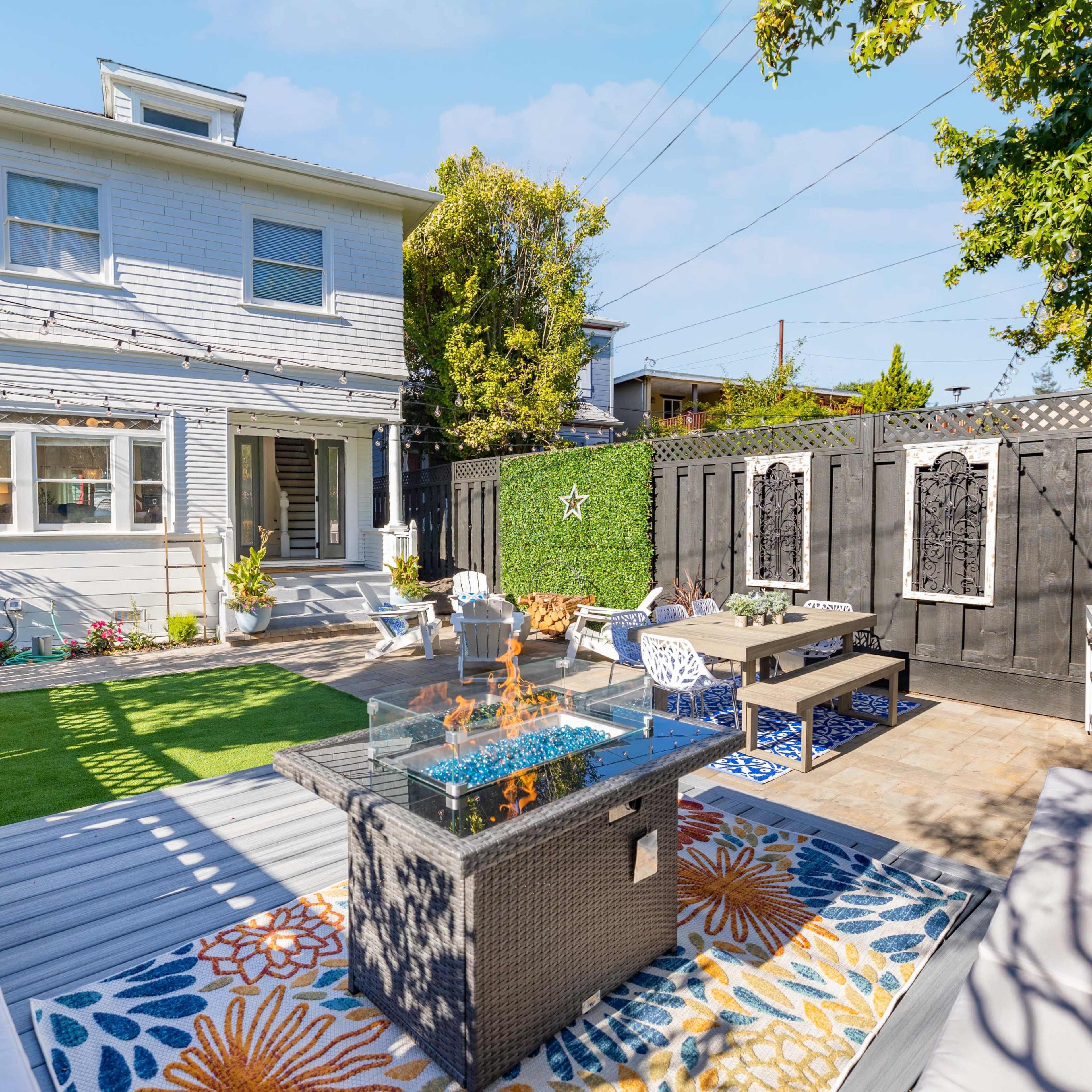 A backyard with a fire pit on a patio surrounded by seating areas, a green lawn, and a two-story house in the background.