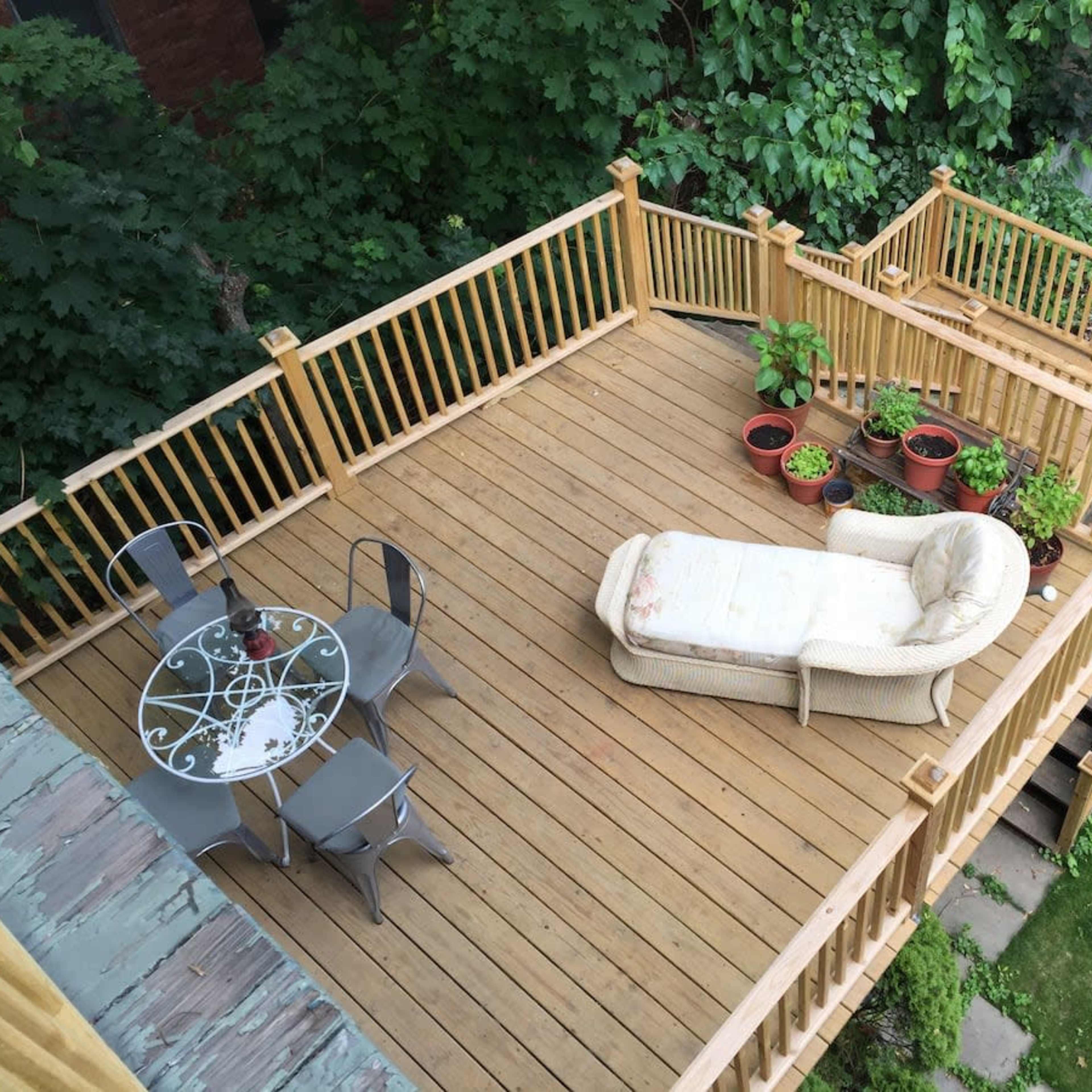 A wooden deck surrounded by greenery, featuring a seating area with a chaise lounge and a small round table with chairs.