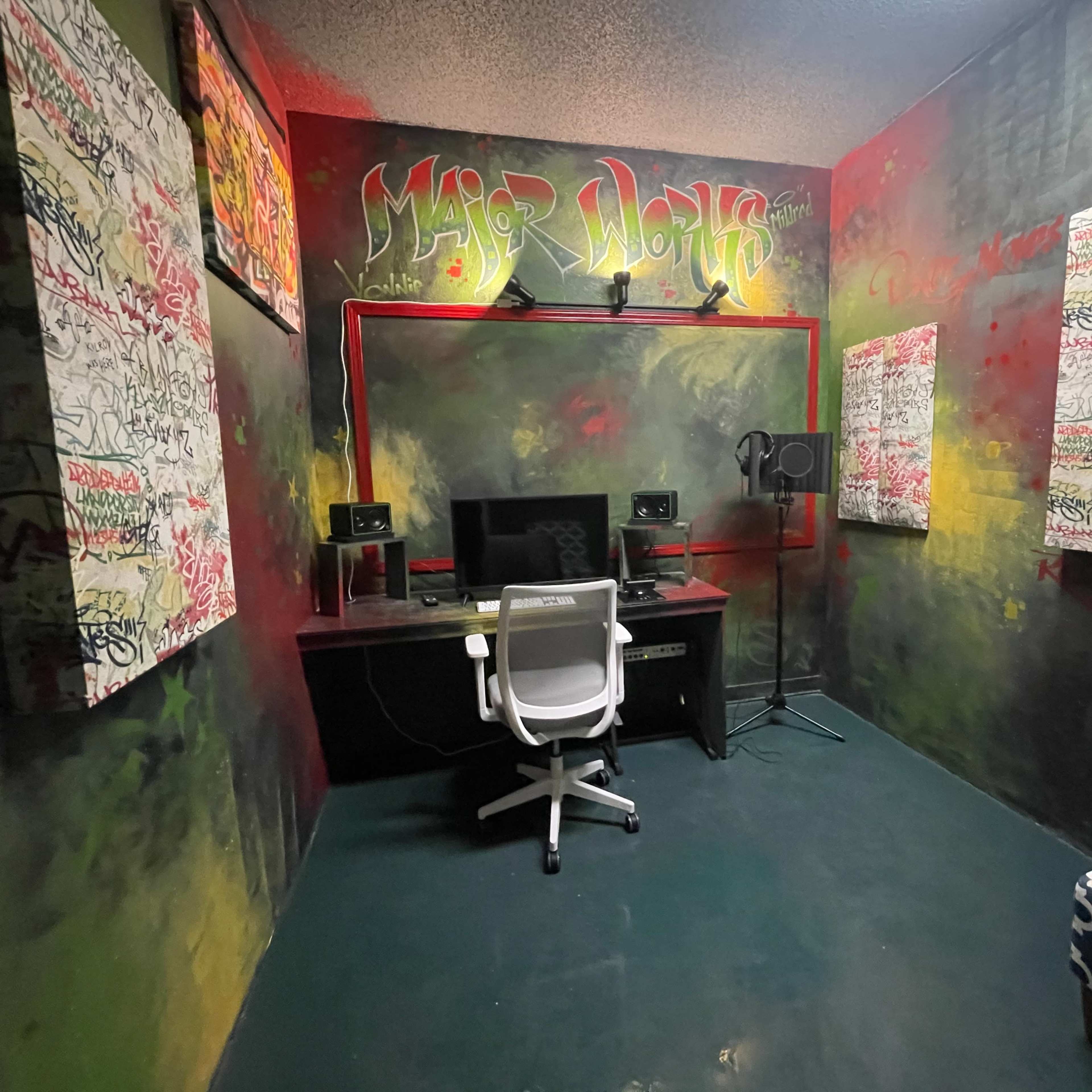 A small recording studio with graffiti-covered walls, a desk with a computer and speakers, a microphone, and a chair.