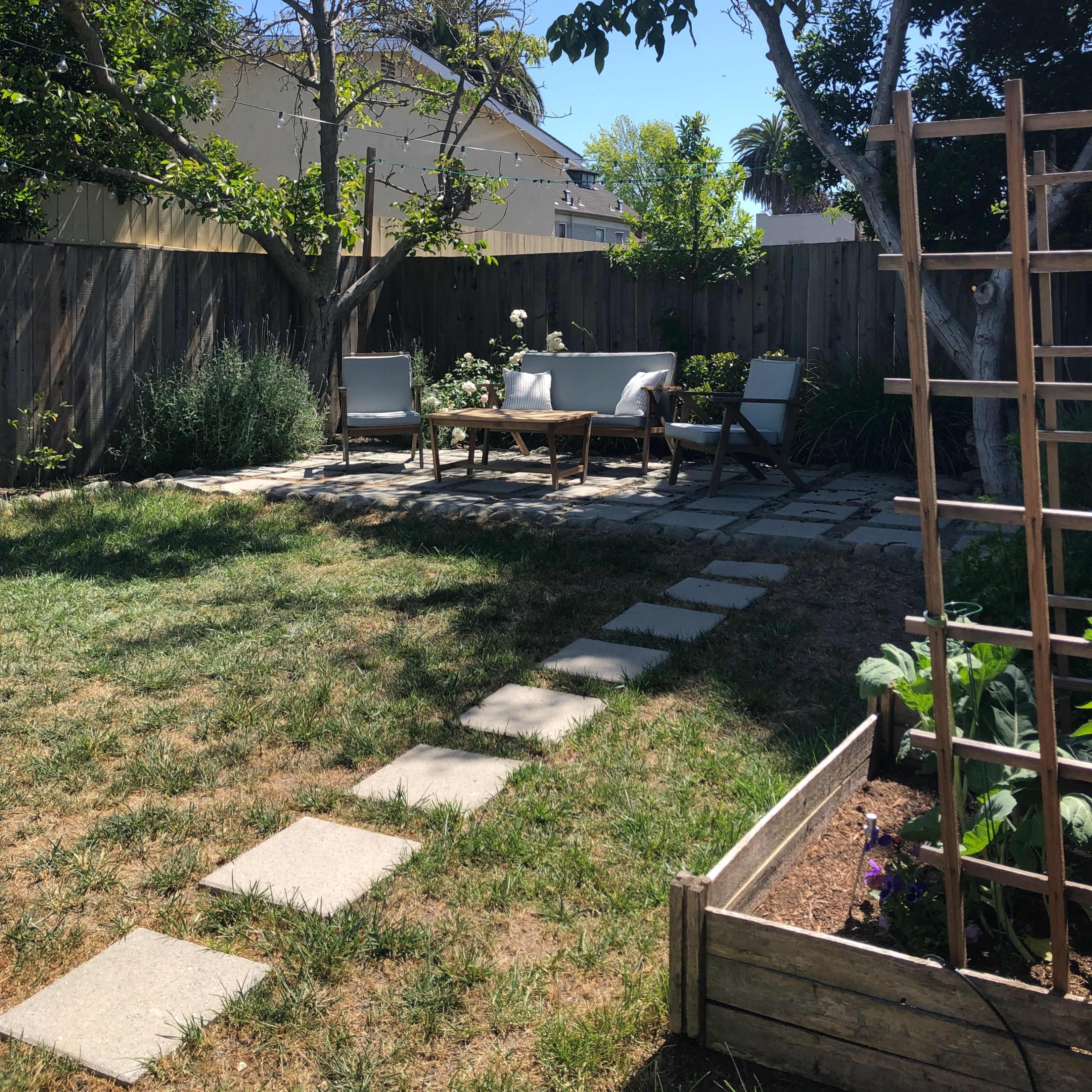 A backyard features a seating area with chairs and a table surrounded by grass and trees, along with a stone path leading to a garden bed.