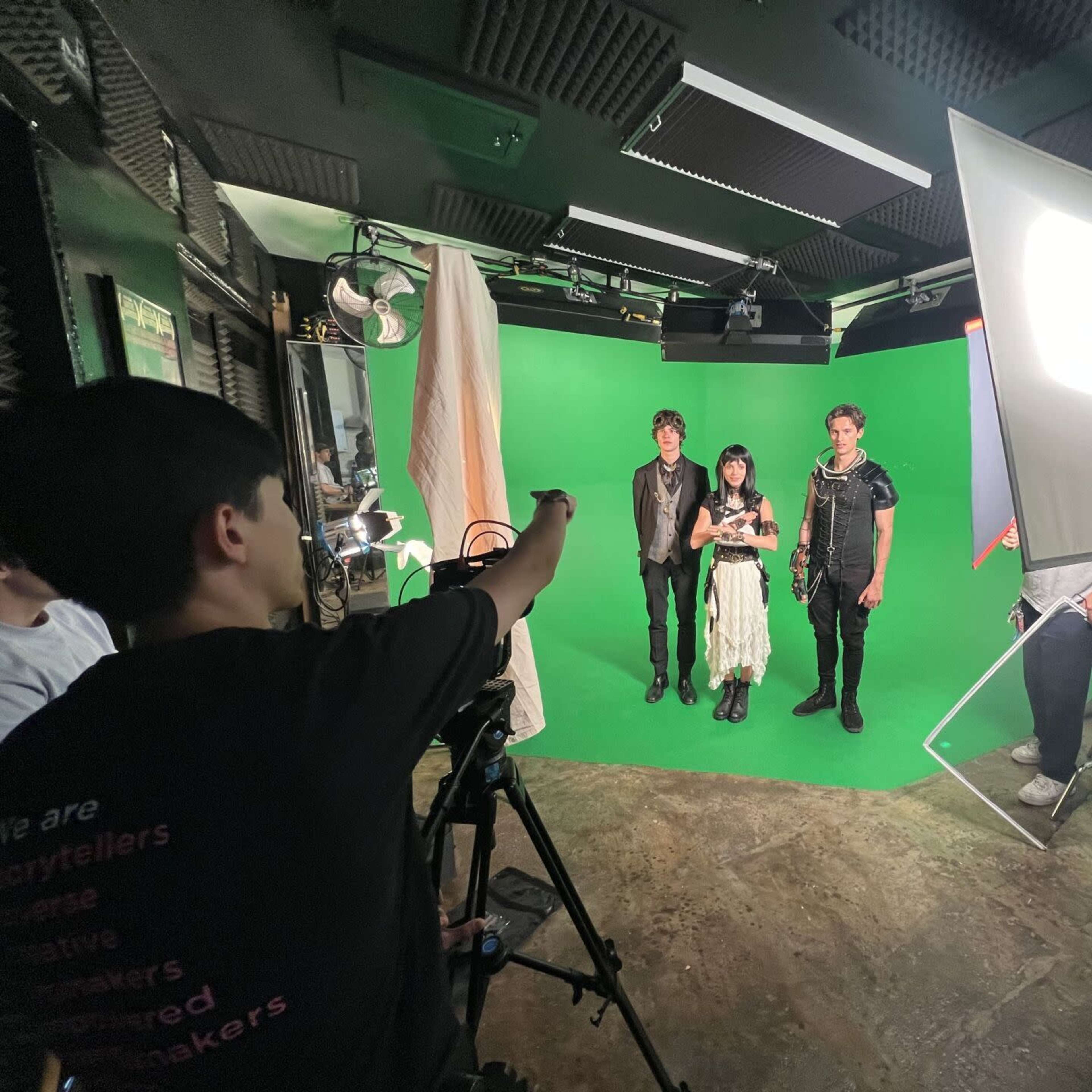 A group of three individuals stands in front of a green screen while a person adjusts equipment nearby.