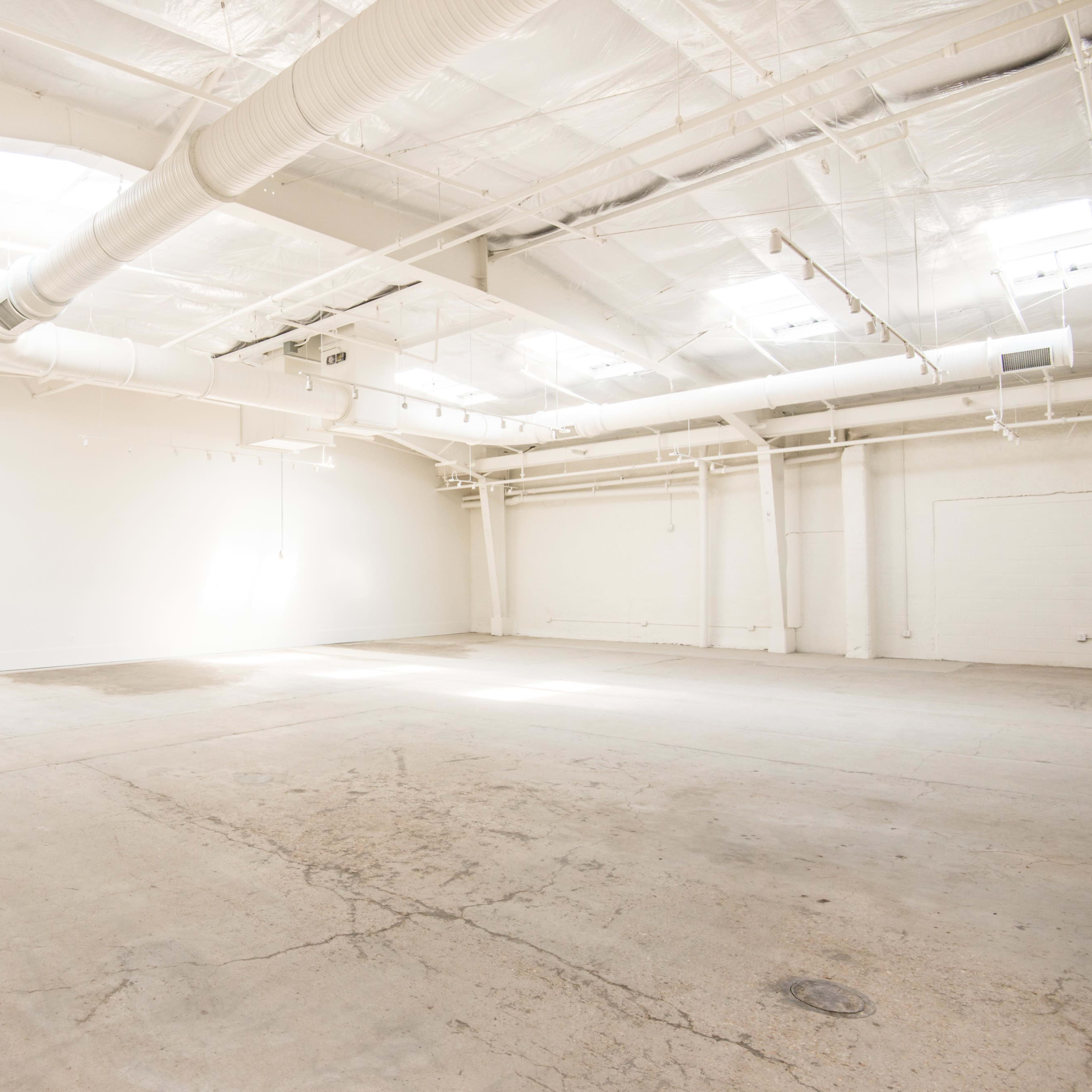 A spacious, empty room with concrete flooring and high ceilings, illuminated by natural light from overhead windows.