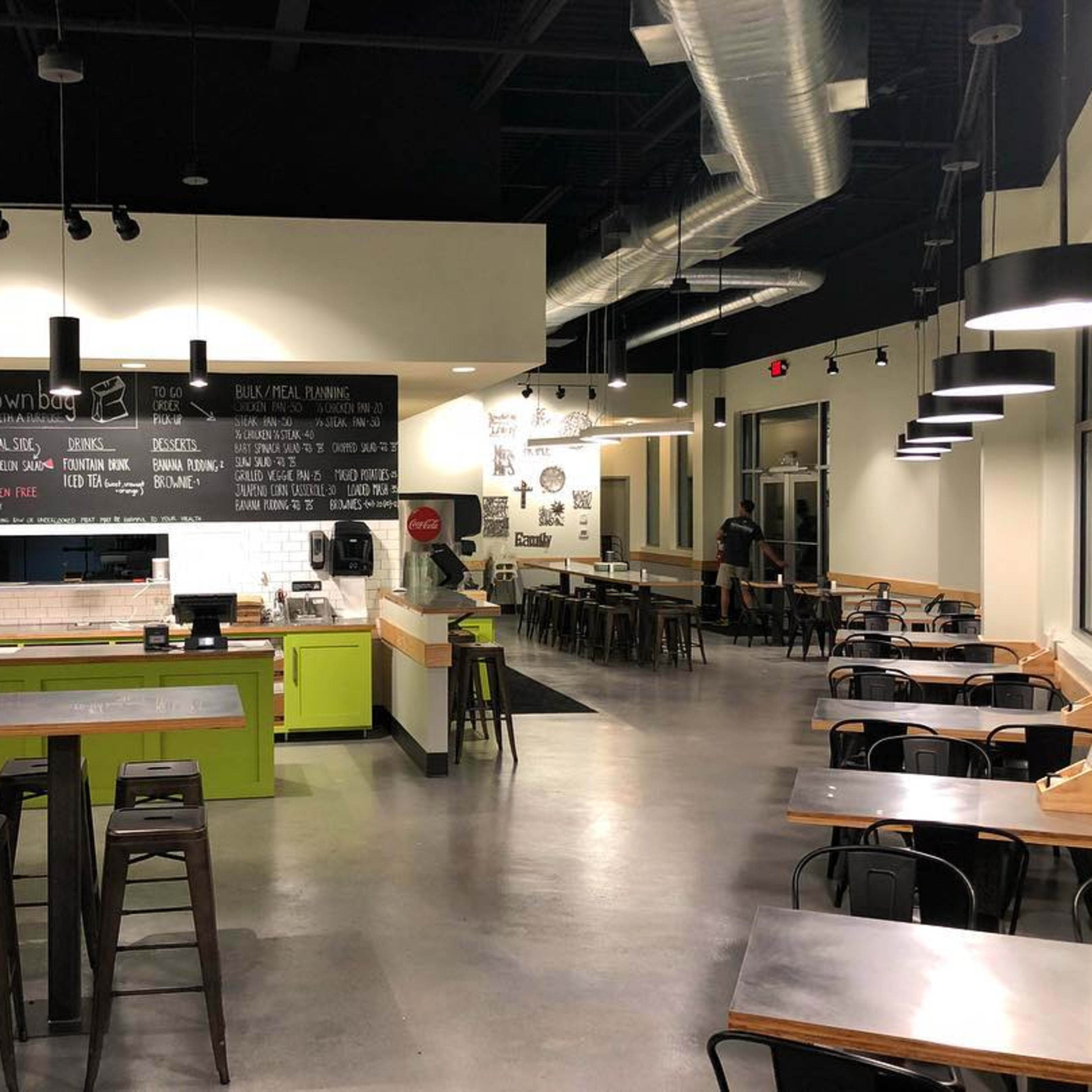 A modern restaurant interior with a green serving area, metal seating, and a chalkboard menu on the wall.