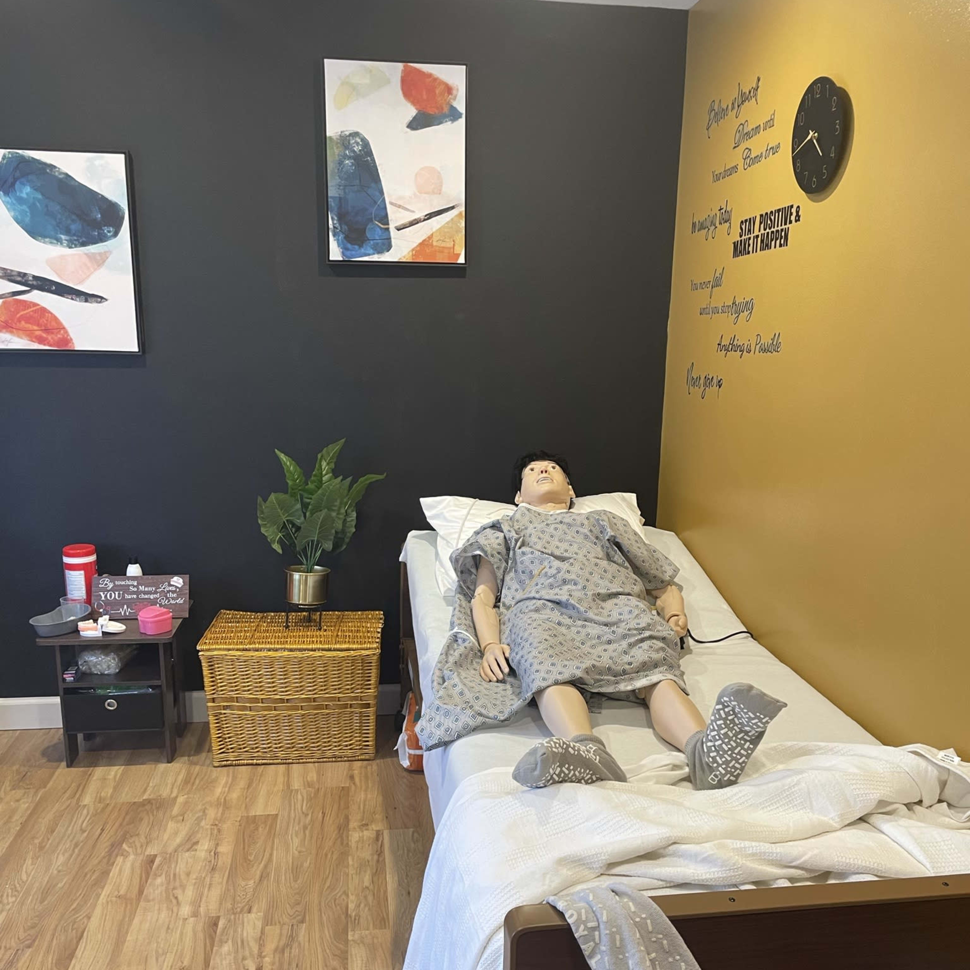 A simulation room with a mannequin lying in a hospital bed, surrounded by a small table, a basket, a plant, and framed artwork on the wall.