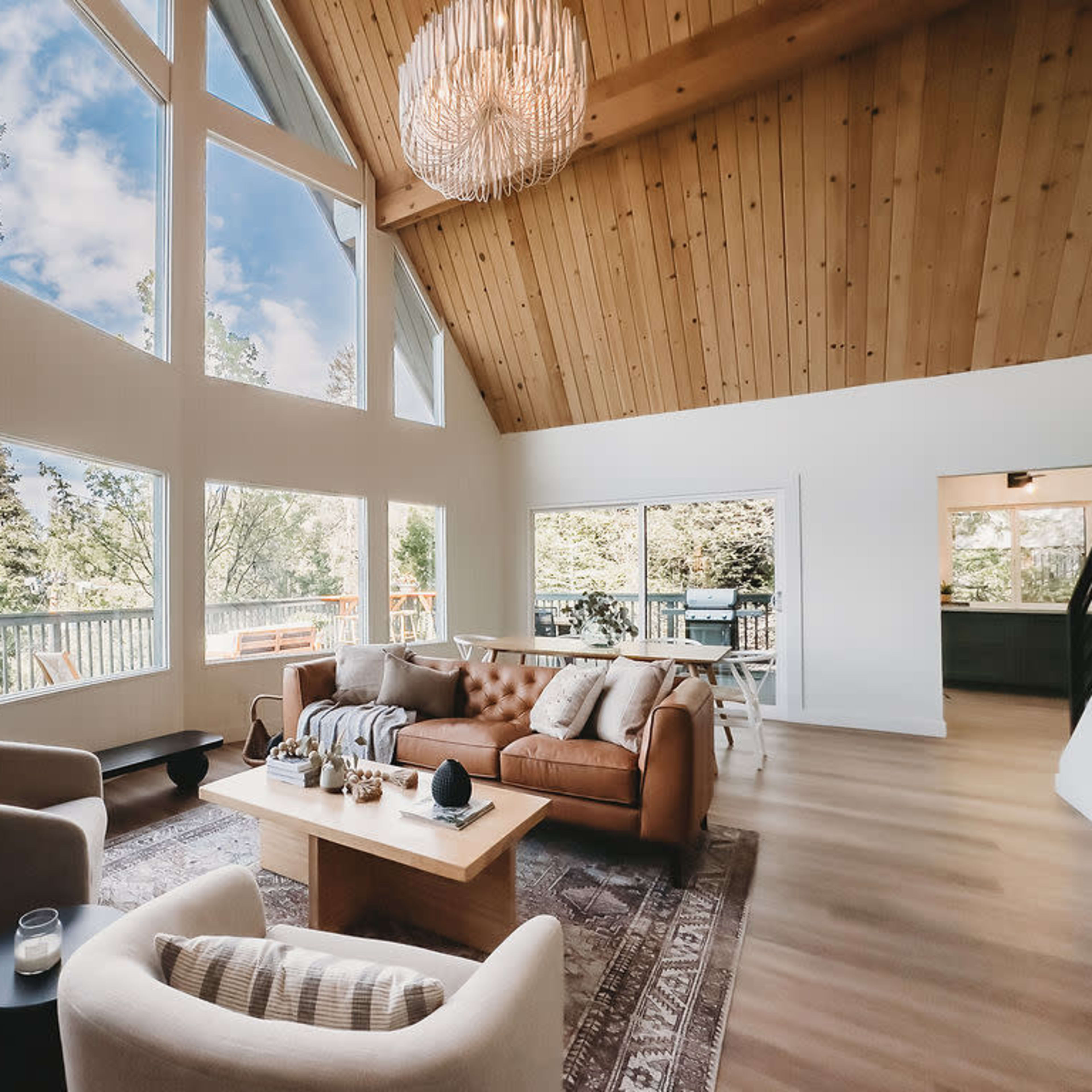 The living room features large windows, a wooden ceiling, modern furniture, and a view of trees outside.
