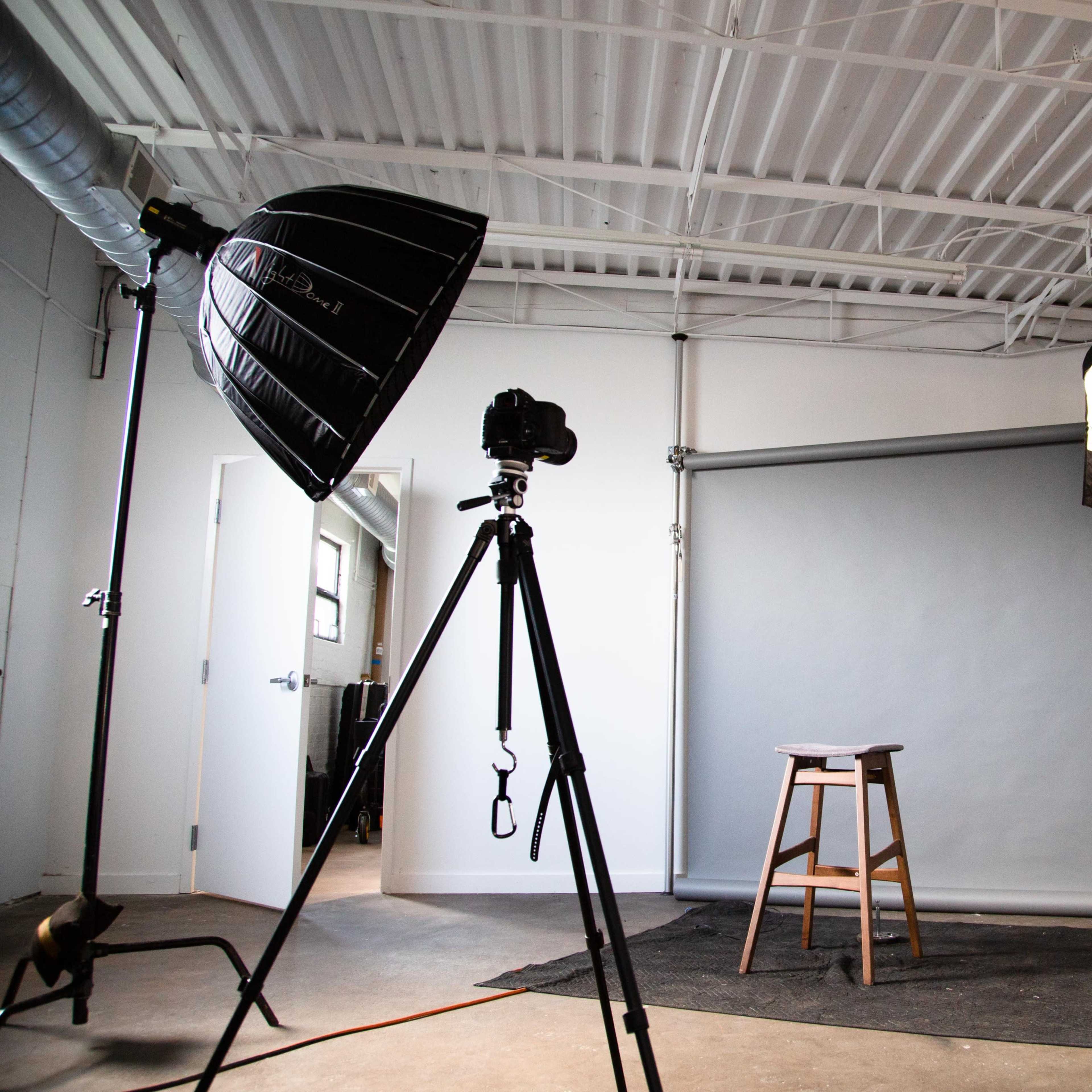 A photography studio with a camera on a tripod, a stool, and lighting equipment set up against a gray backdrop.
