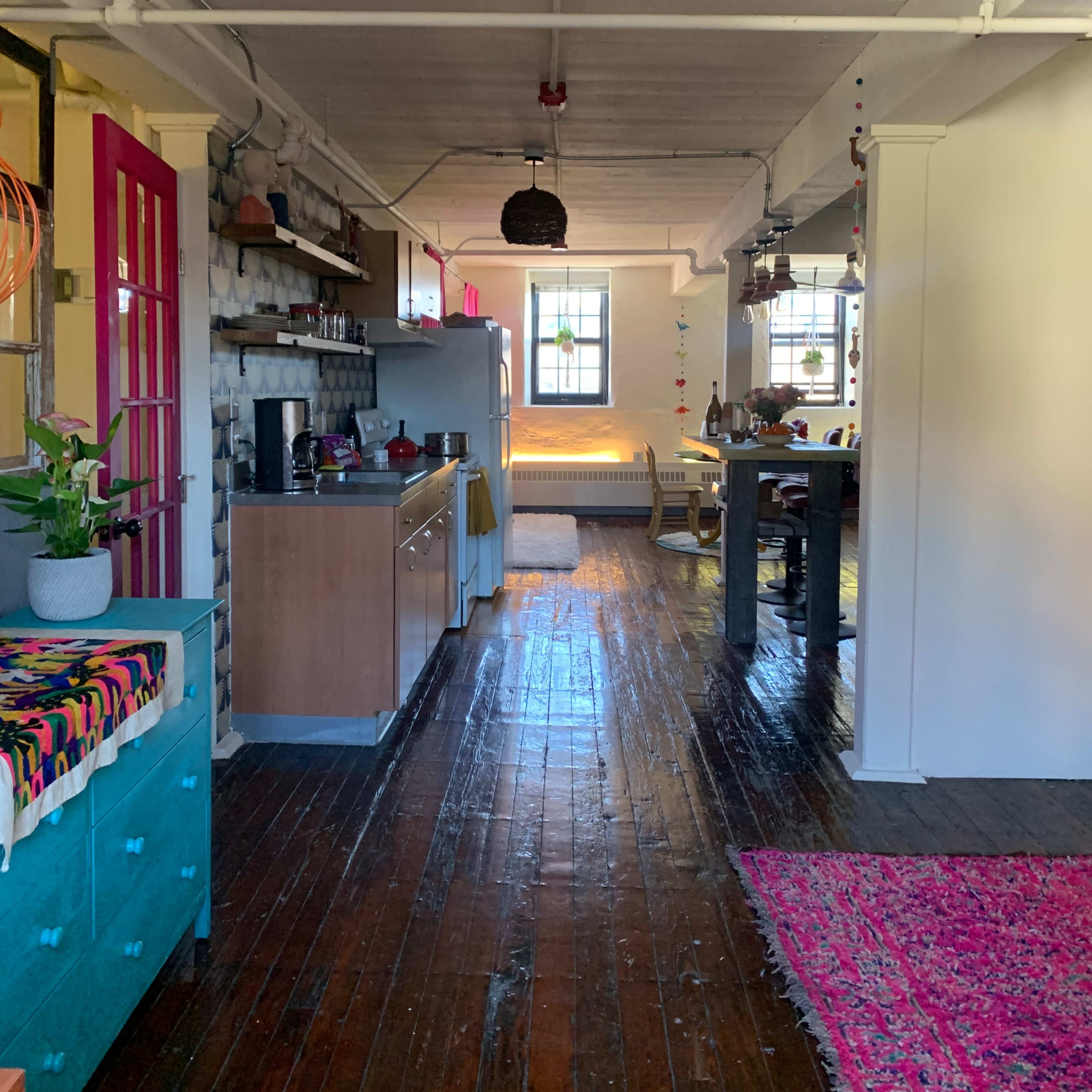 A long, narrow kitchen and dining area with wooden floors, featuring a mix of colorful cabinets, a stainless steel refrigerator, and a dining table set against a bright backdrop of windows.