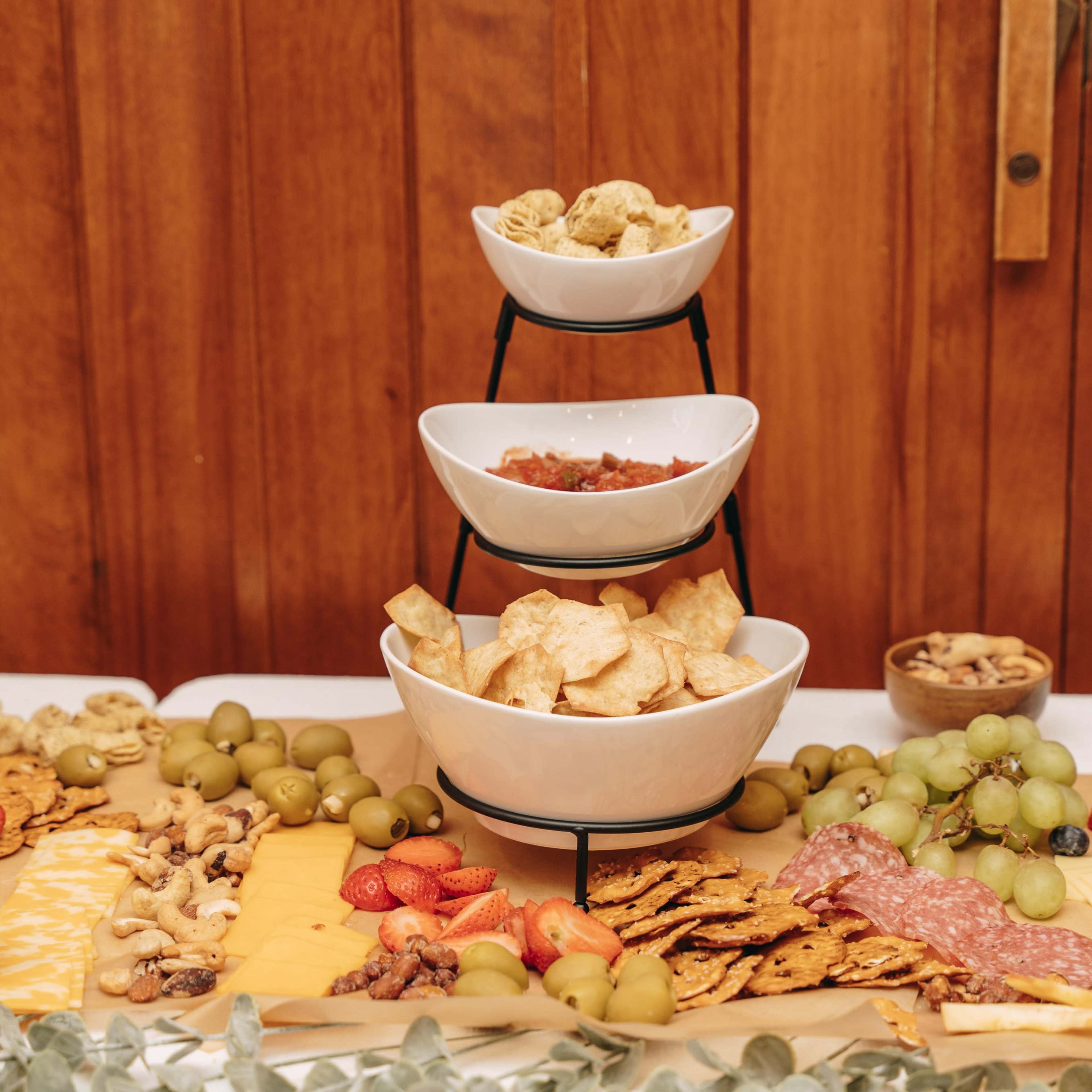 A tiered serving stand displays bowls of snacks, including chips and salsa, surrounded by an assortment of fruits, nuts, and crackers on a table.