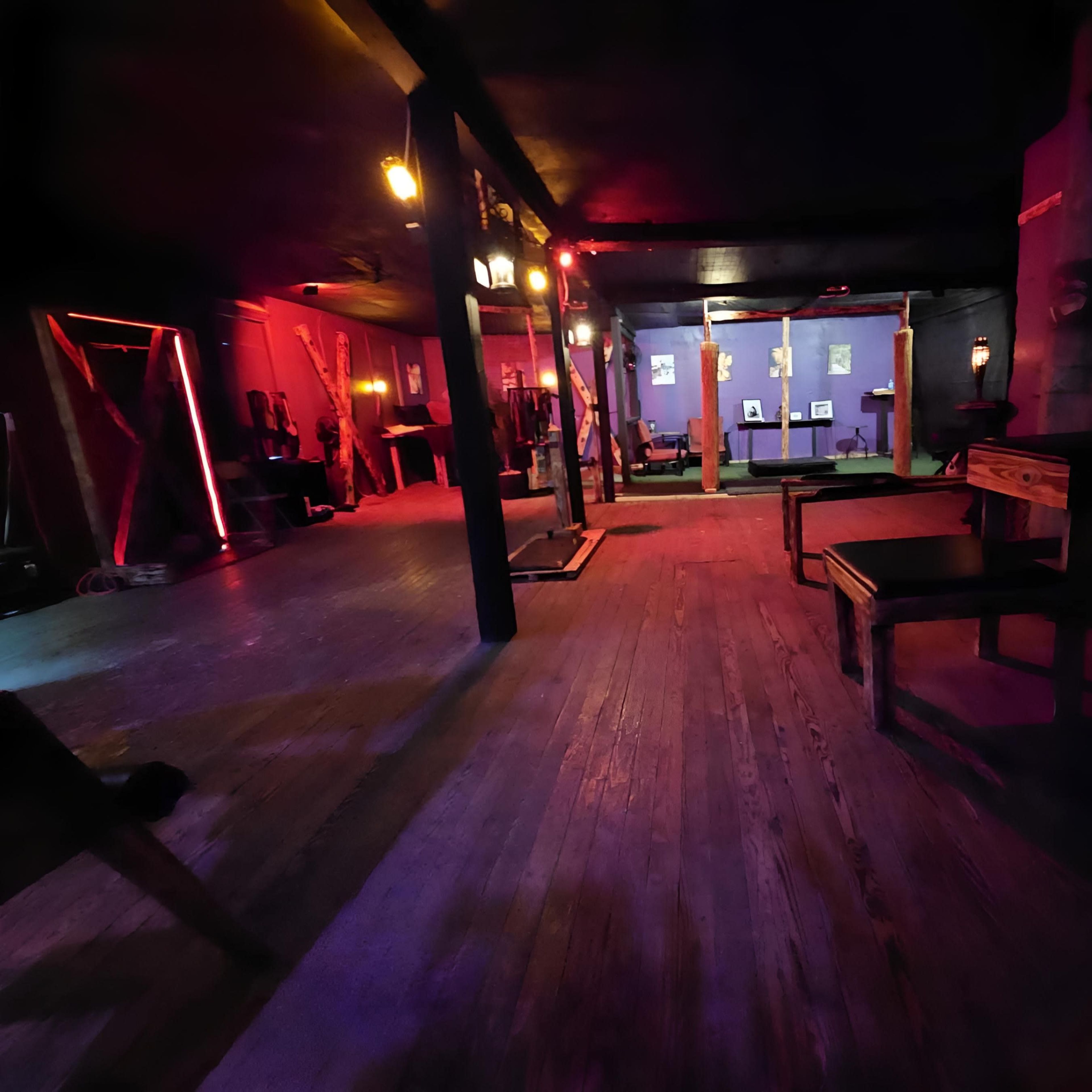 A dimly lit interior with wooden flooring, tables, and colorful lighting effects creating a moody atmosphere.