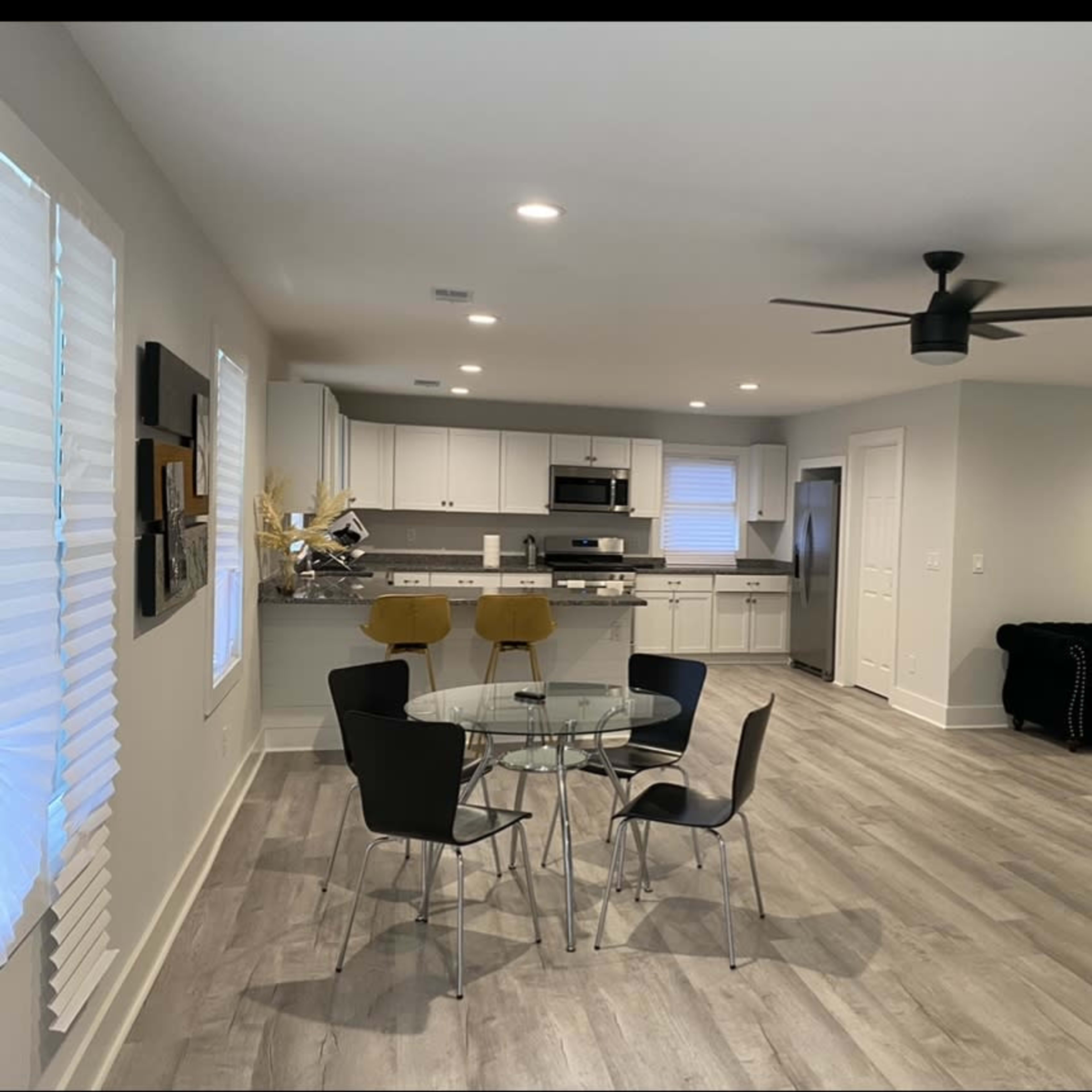 A modern, open-concept living space with a dining area featuring a glass table and four chairs, a kitchen with white cabinetry, and a living area with a black sofa.