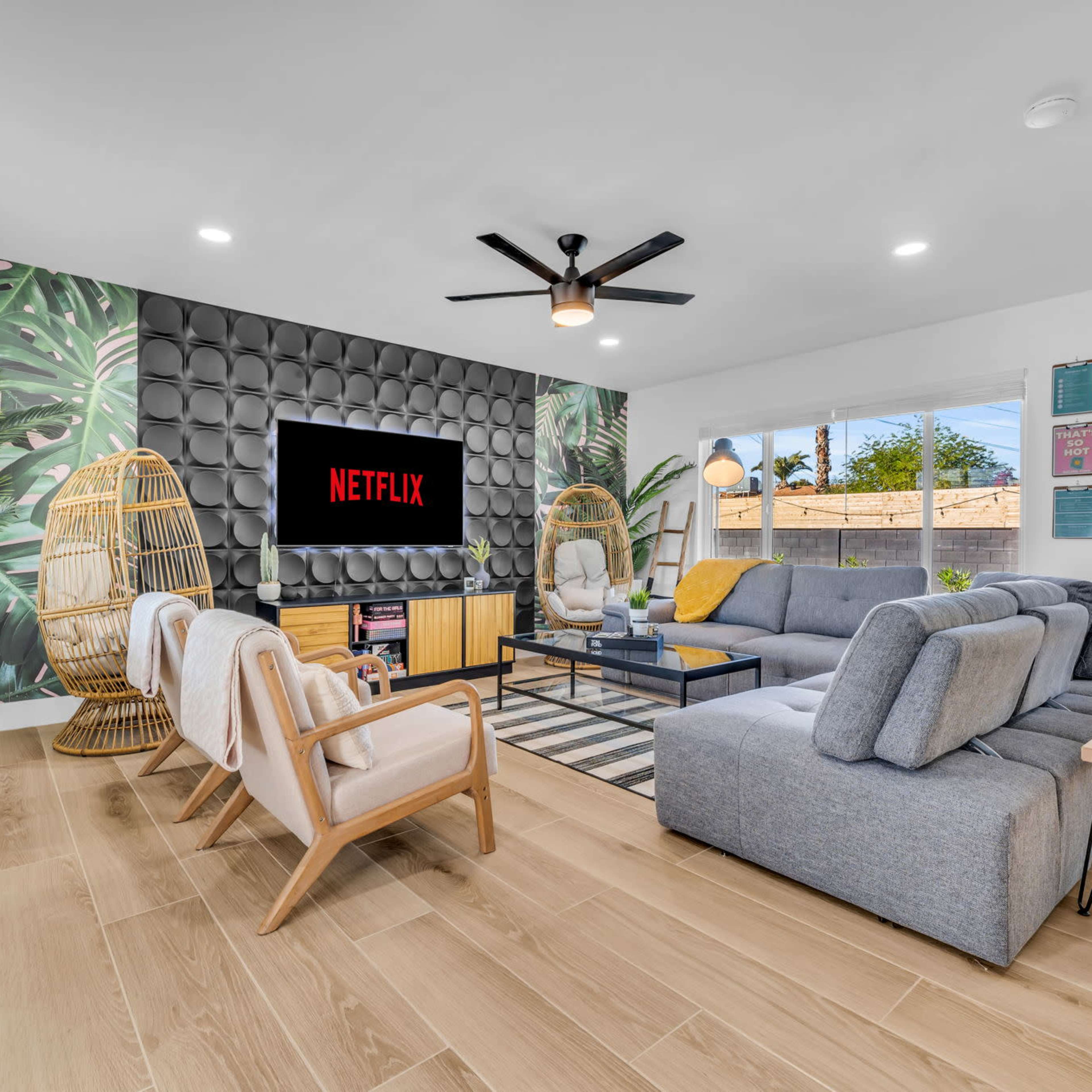 A modern living room features a large gray sectional sofa, two woven chairs, and a wall-mounted TV displaying the Netflix logo, set against a backdrop of tropical-themed wallpaper.