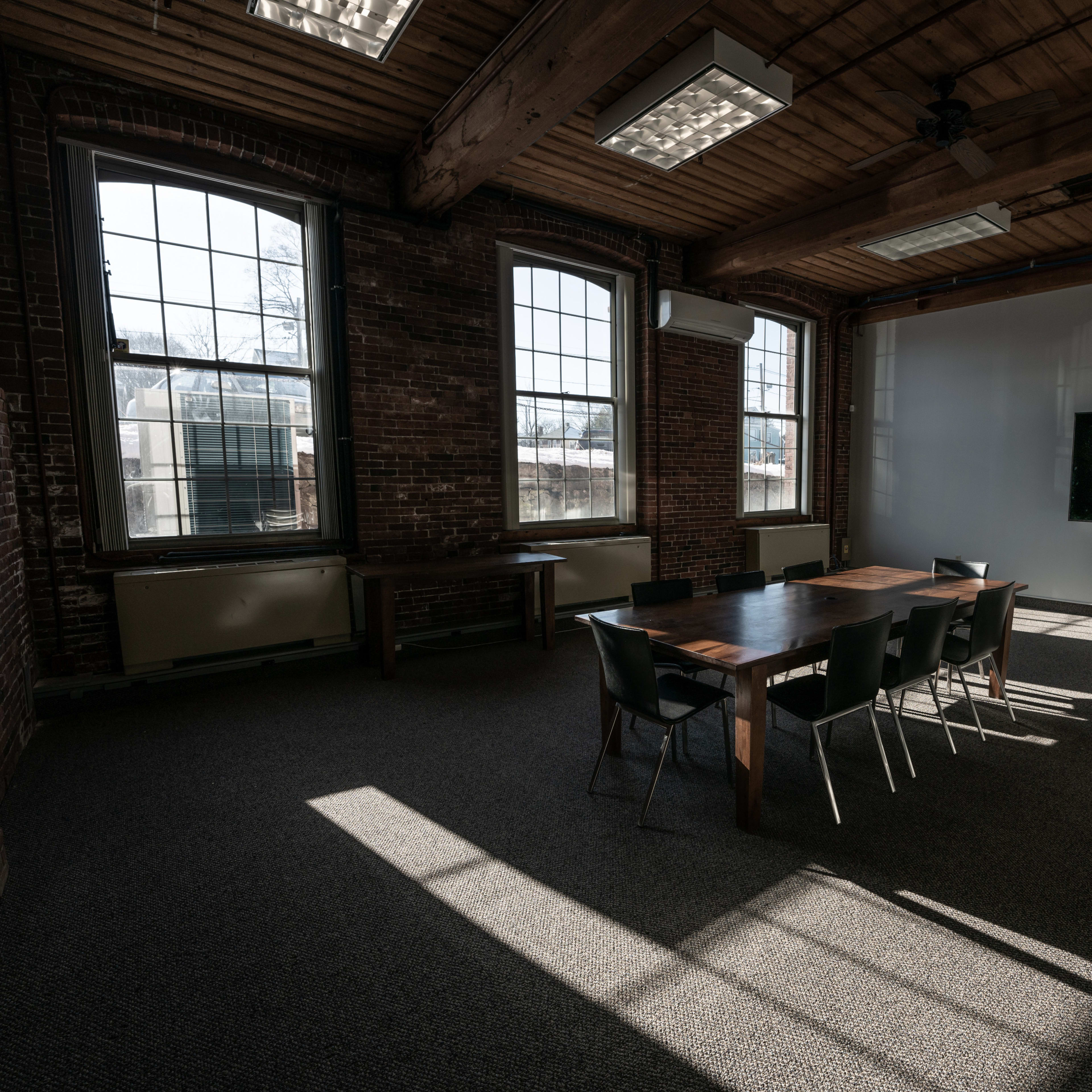 A spacious meeting room features a large wooden table surrounded by chairs, with large windows allowing light to cast shadows on the carpeted floor.