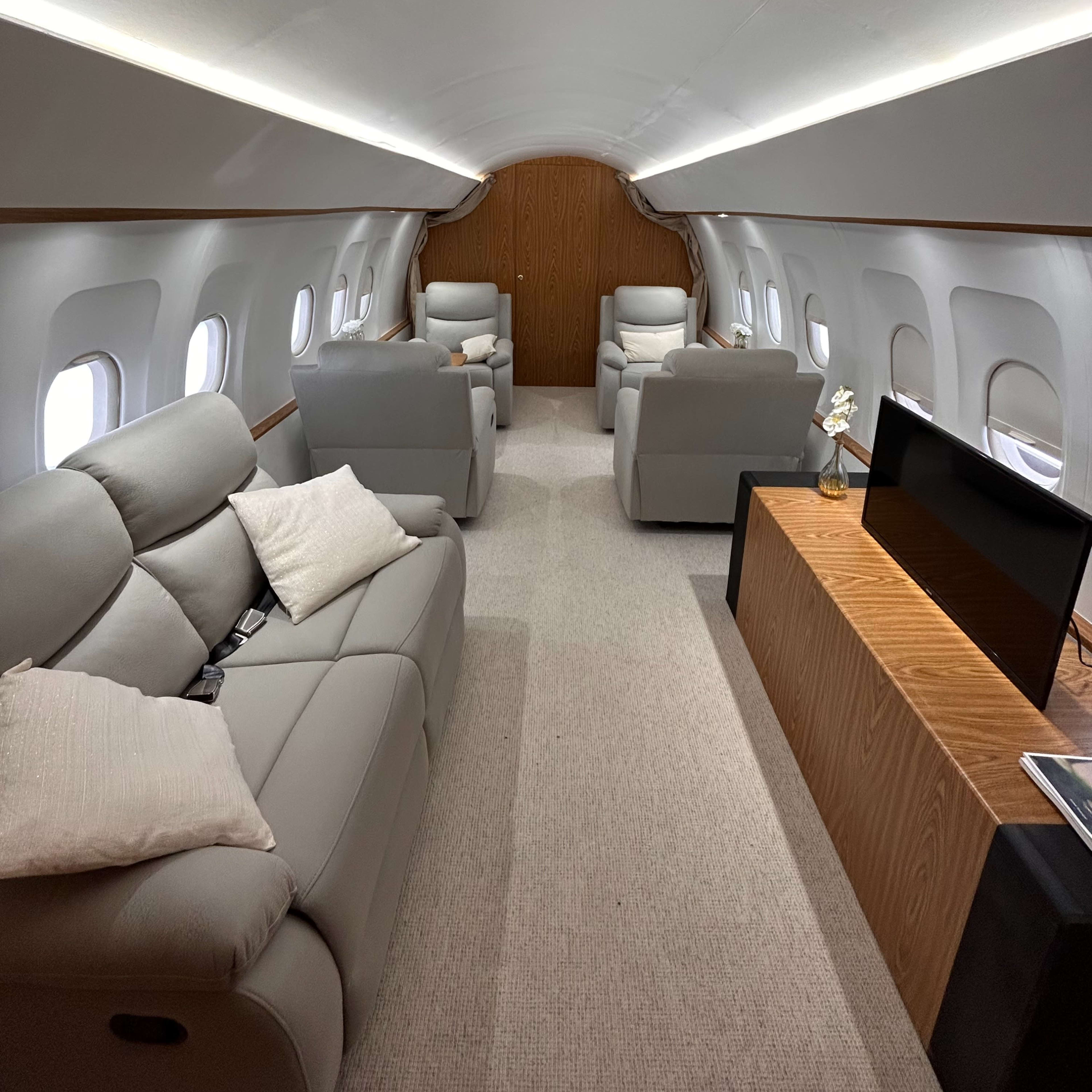 The interior of a private jet features two light gray sofas, several armchairs, and a wooden cabinet along the side, with large windows allowing natural light to illuminate the space.