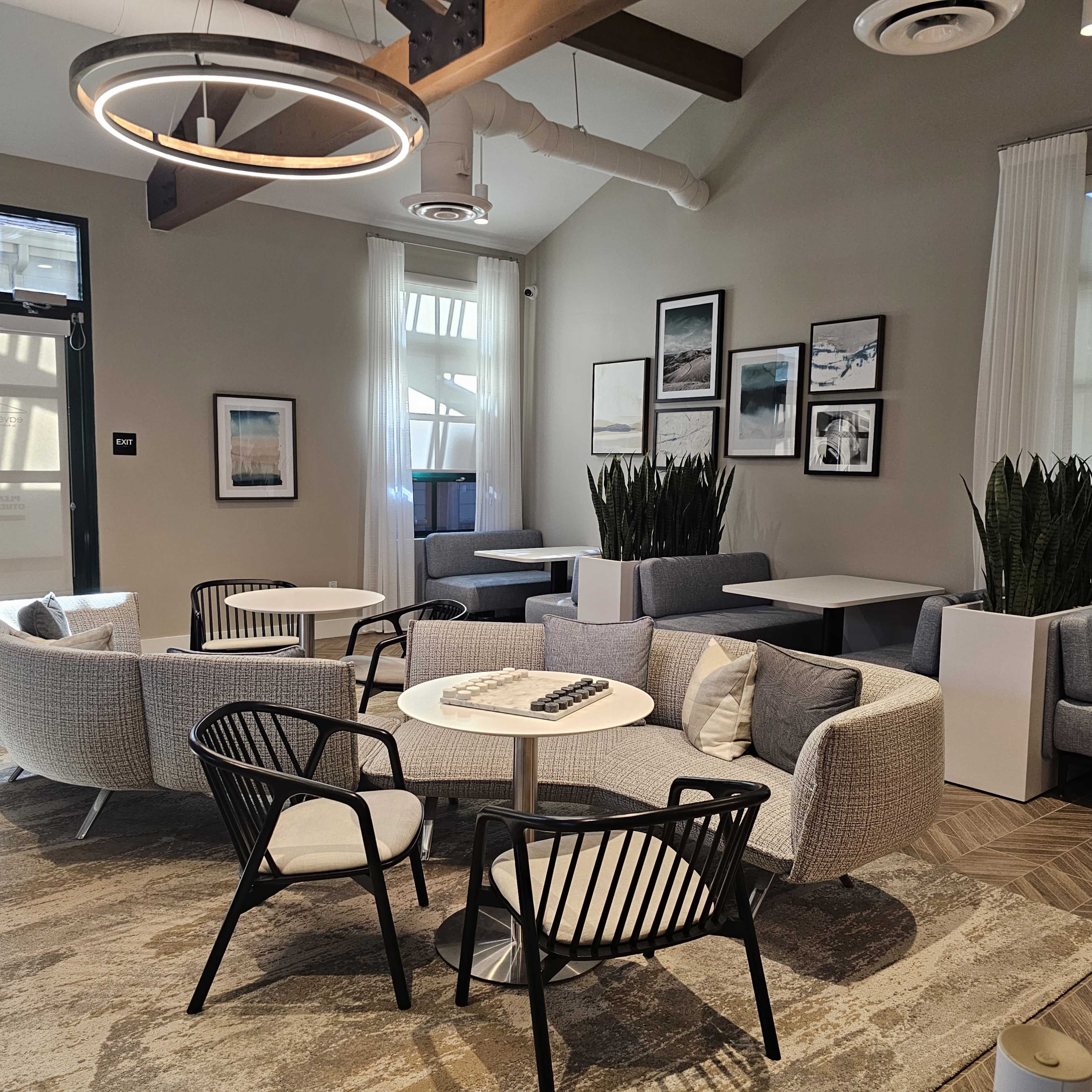 A modern lounge area with multiple seating arrangements, including circular tables and a mix of chairs, framed artwork on the walls, and large plants.