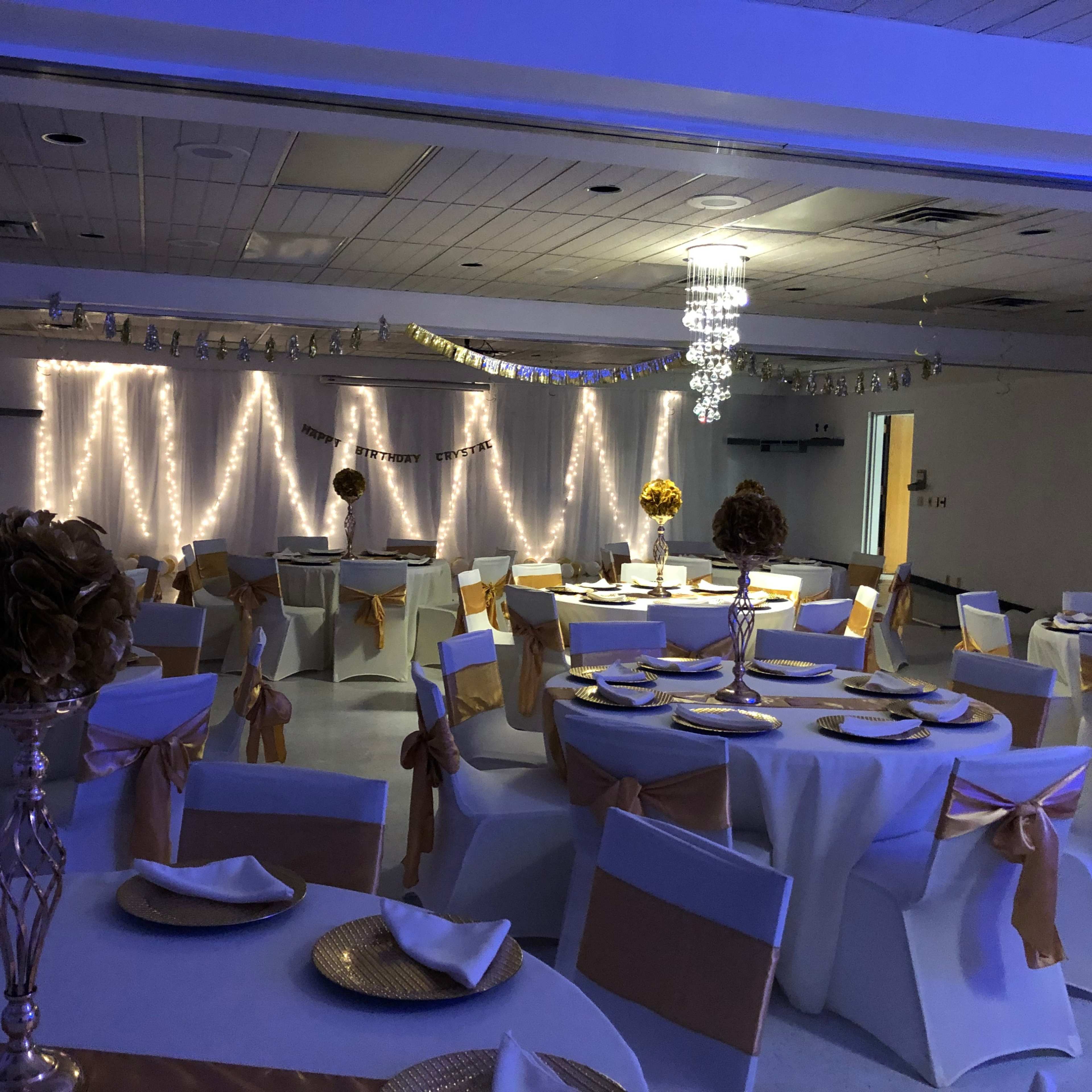 The interior of a banquet hall is set with round tables covered in white tablecloths and adorned with gold accents, illuminated by blue lighting and decorative string lights.