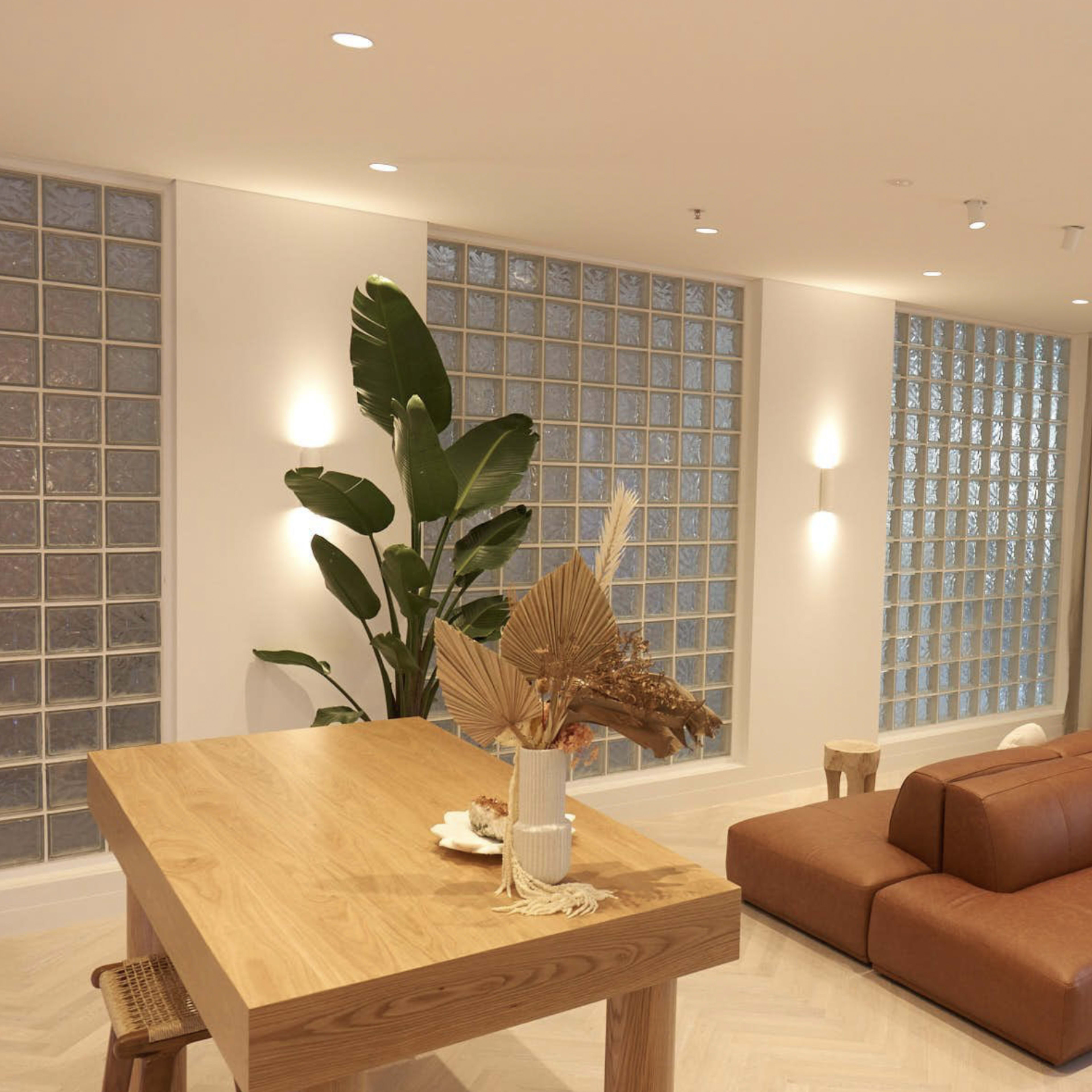 A modern interior space featuring a brown leather sofa, a wooden dining table, large glass block windows, and a circular mirror on the wall.