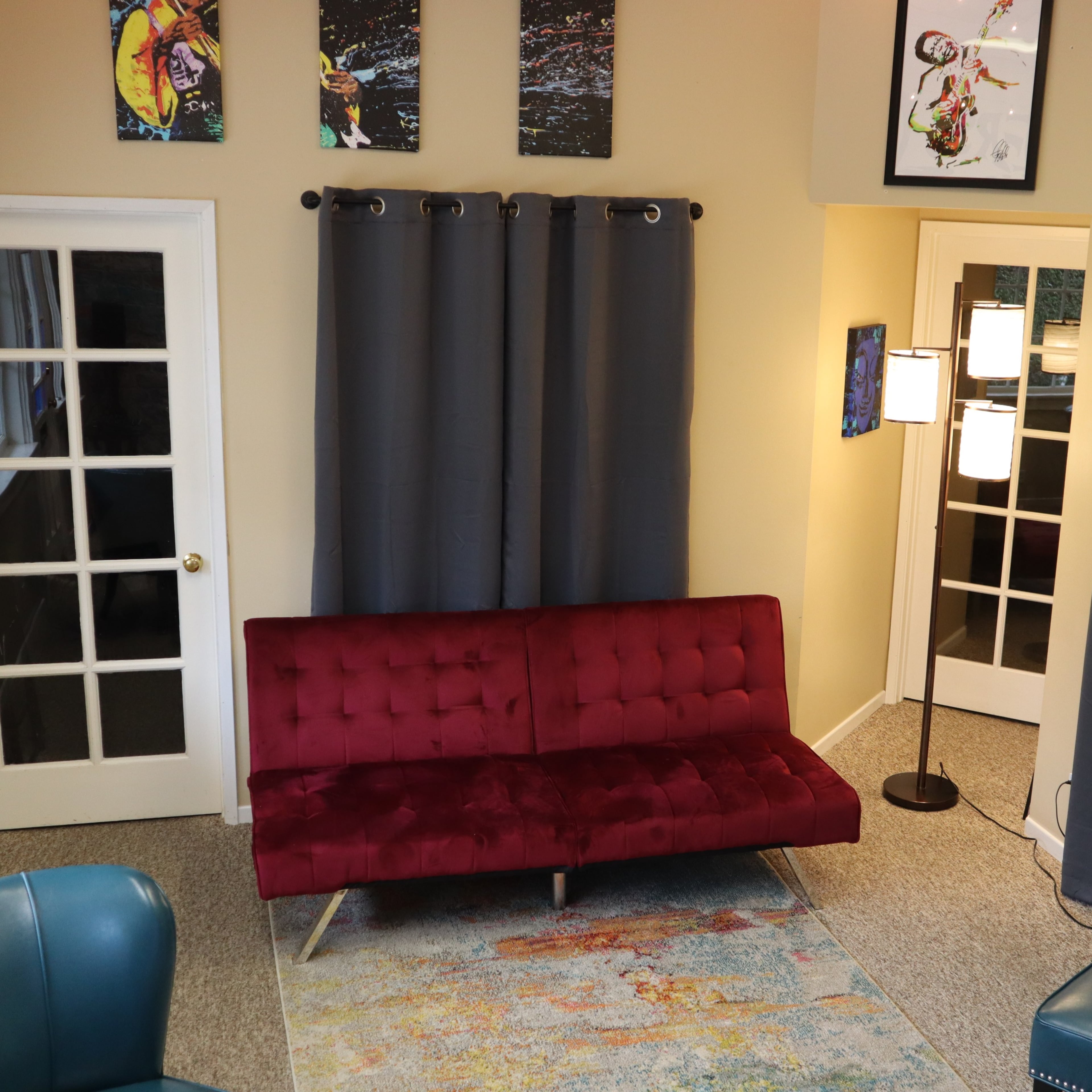 The room features a red velvet couch, two teal chairs, gray curtains, and artwork on the walls, with a floor lamp in one corner.