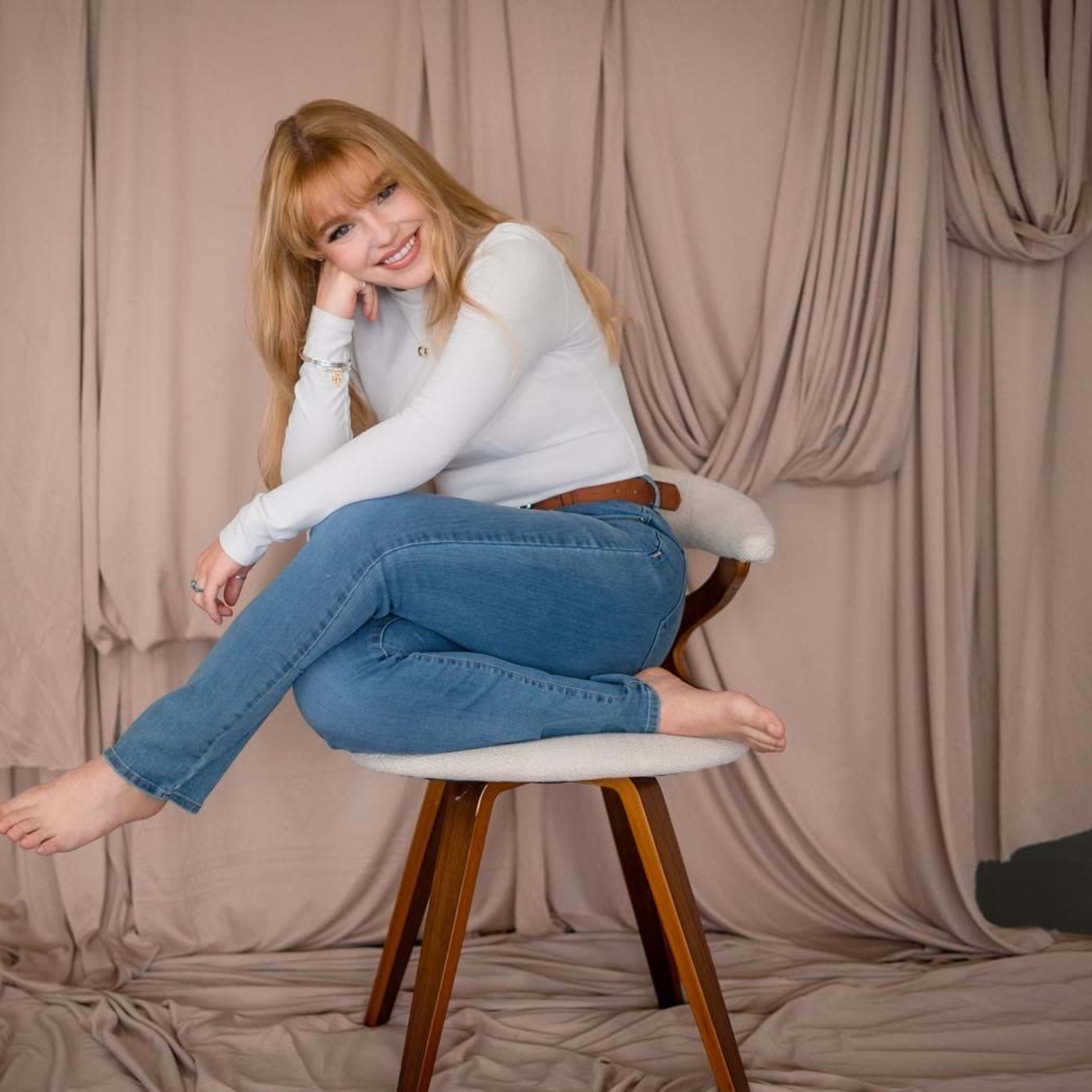 A person with long blonde hair sits on a wooden chair against a background of draped fabric.