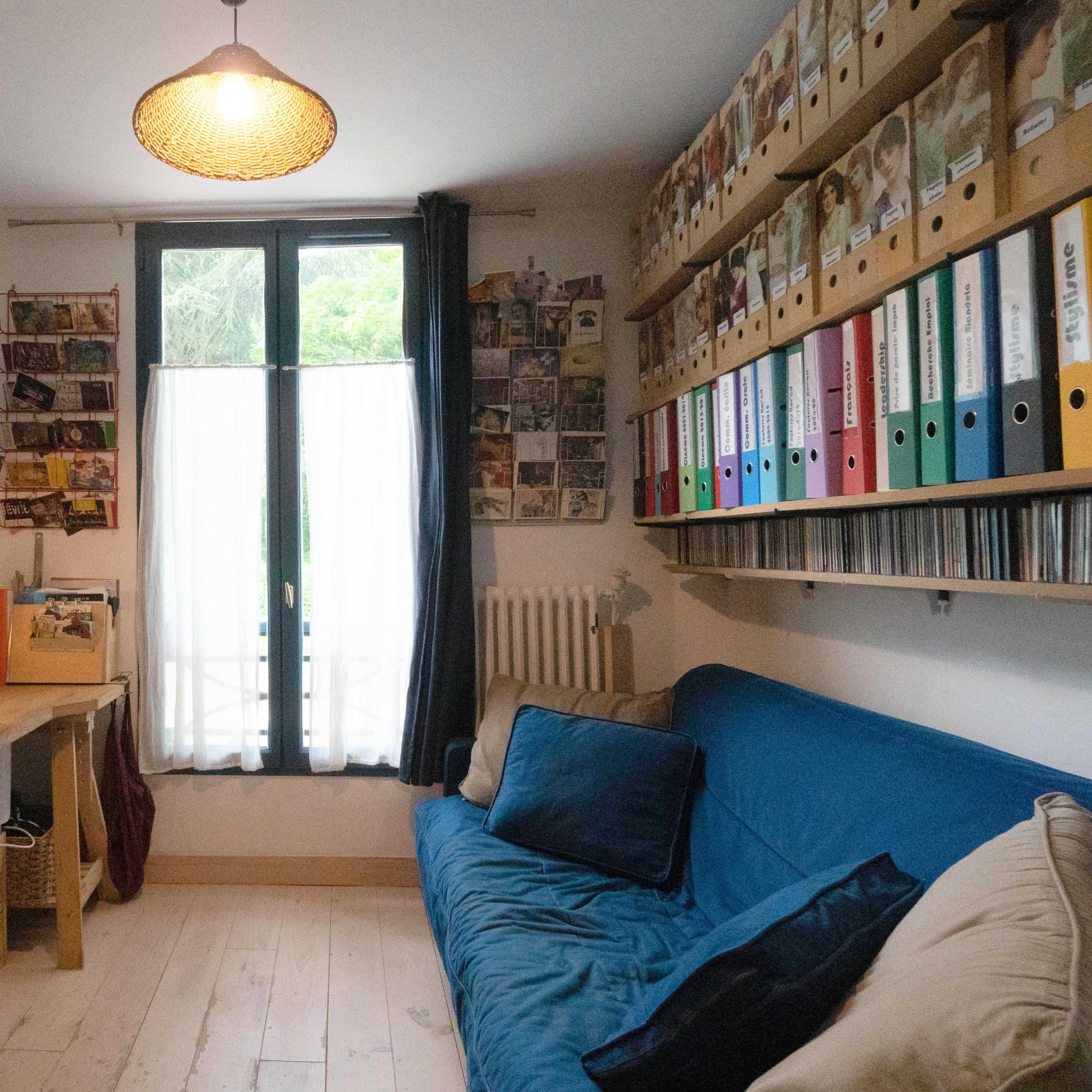 A home office features a blue sofa, a desk with a computer, and shelves filled with colorful binders and photo frames.