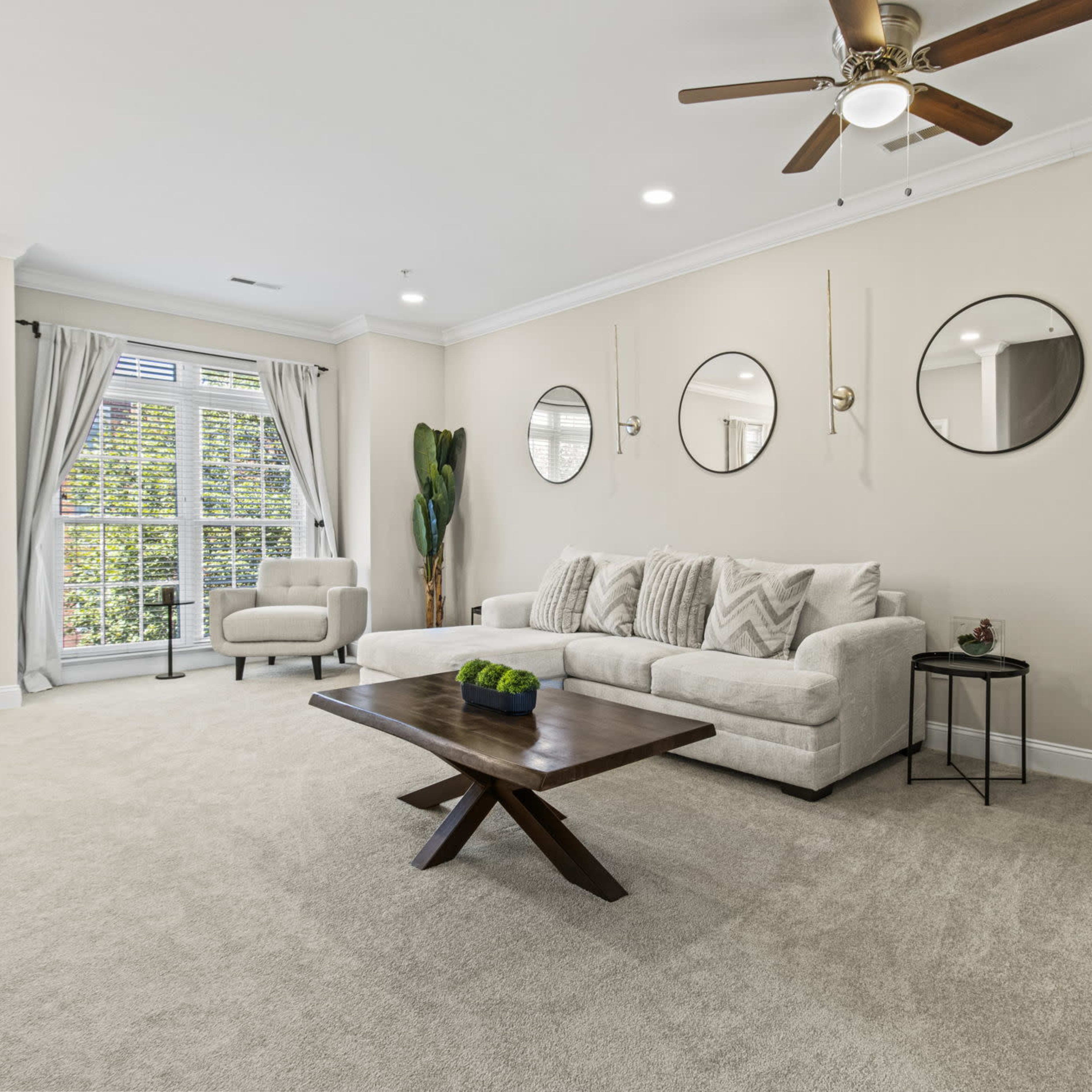 A light-filled living room features a beige sofa, a wooden coffee table, and circular mirrors on the wall, with large windows framed by curtains.