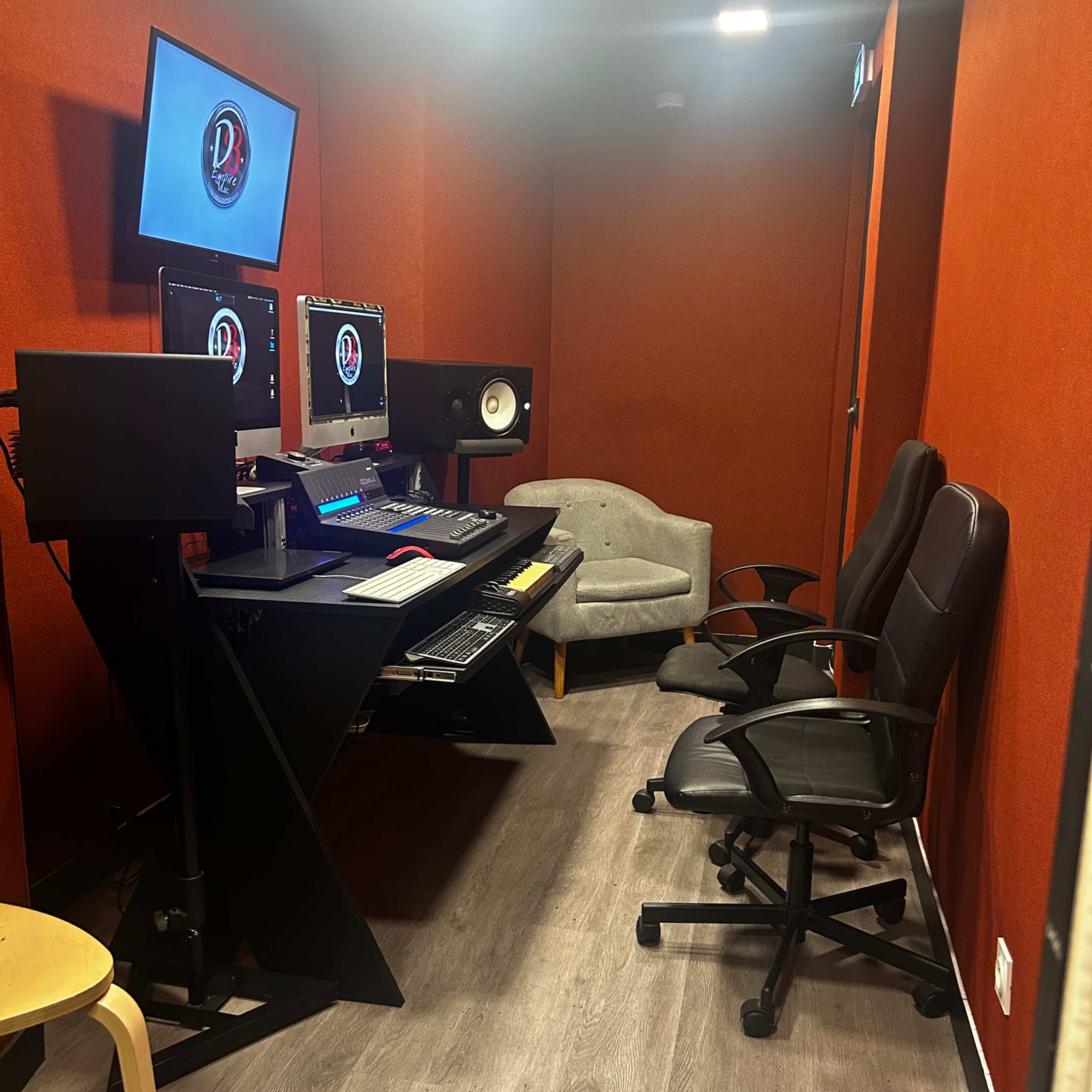 A small recording studio with a black desk featuring multiple computer monitors, speakers, and two office chairs against a bright orange wall.