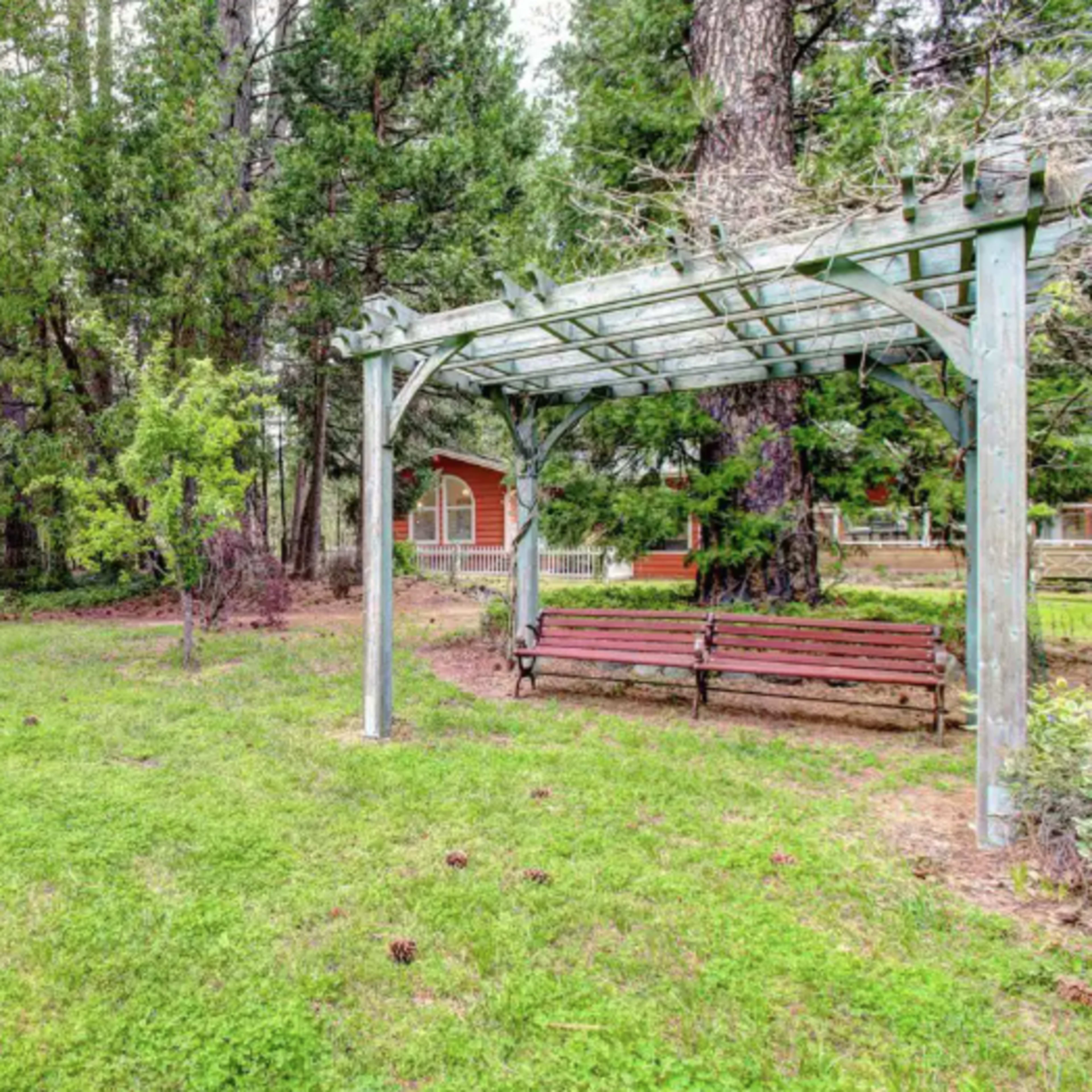 A grassy area with a wooden bench under a pergola, surrounded by trees and a rustic vehicle in the background.