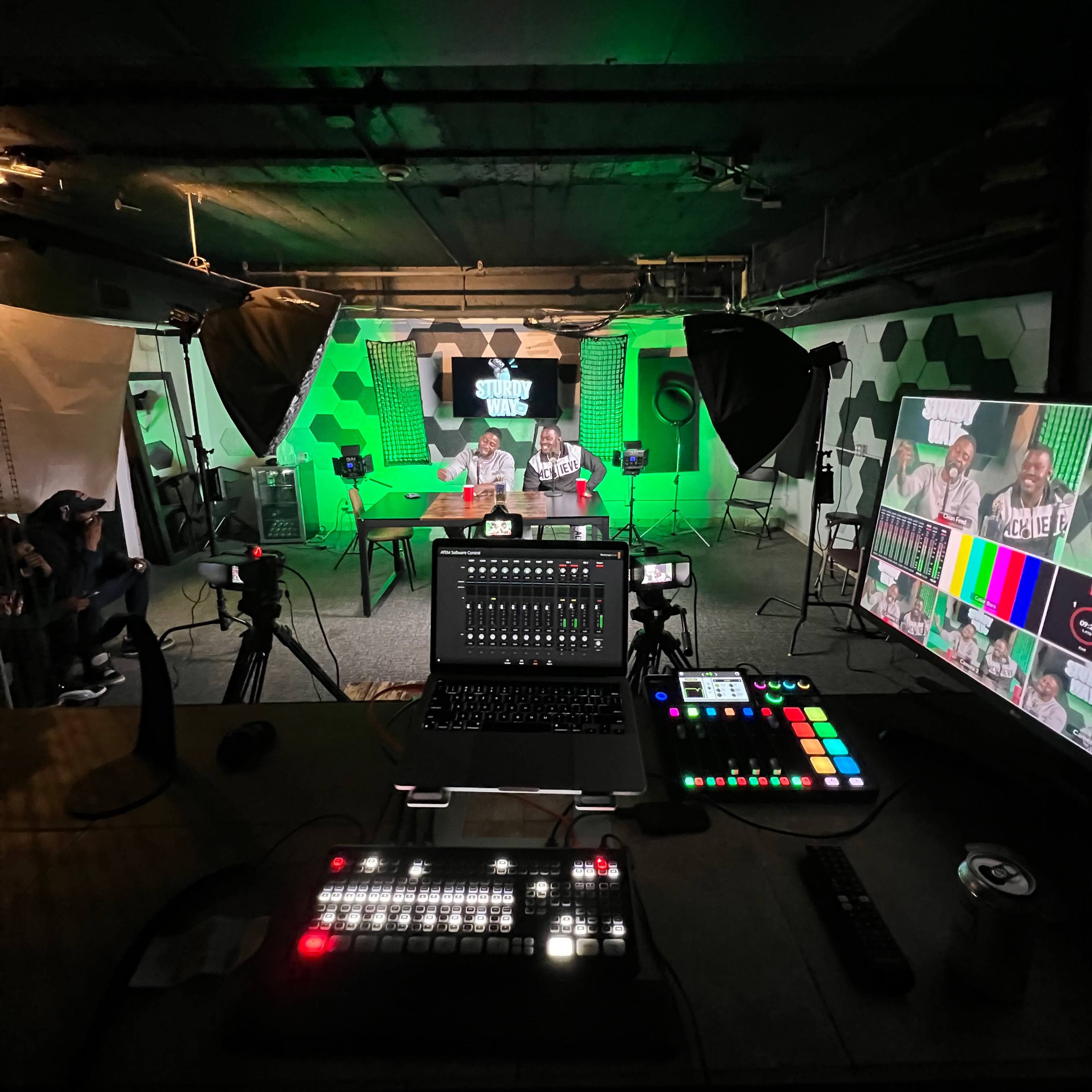A recording studio setup with two people seated at a table under green lighting, surrounded by cameras, monitors, and production equipment.